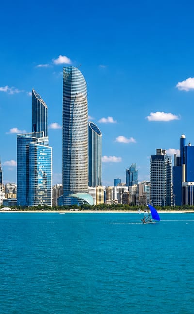 Abu Dhabi's Corniche skyline of glass towers above the turquoise Gulf with sailboats and a UAE flag.