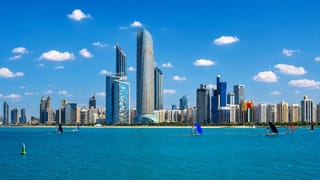 Abu Dhabi's Corniche skyline of glass towers above the turquoise Gulf with sailboats and a UAE flag.
