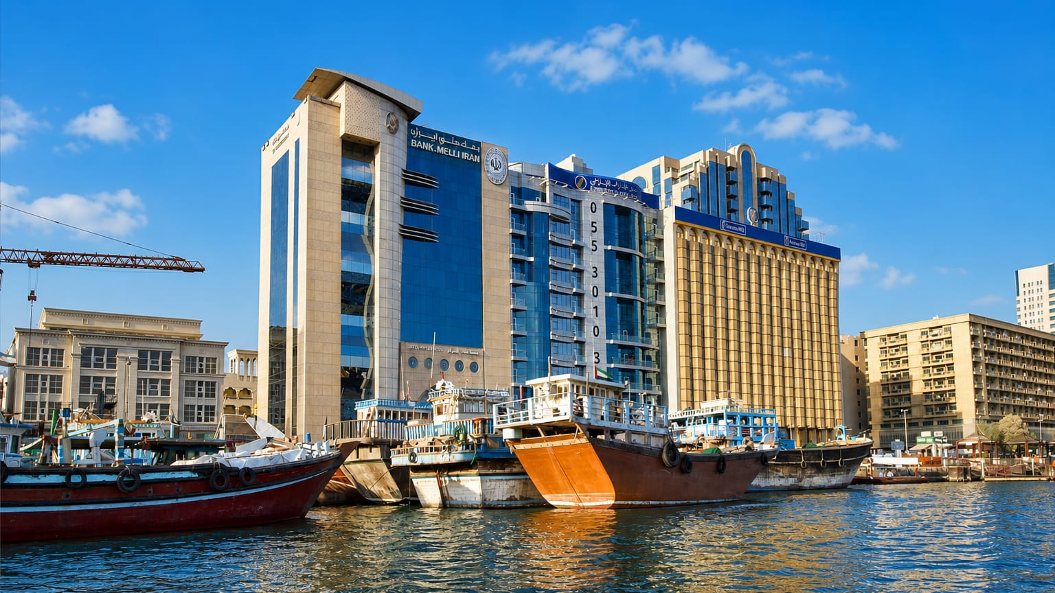 Traditional wooden dhow trading boats moored along Dubai Creek in Deira, with mid-rise commercial buildings behind.