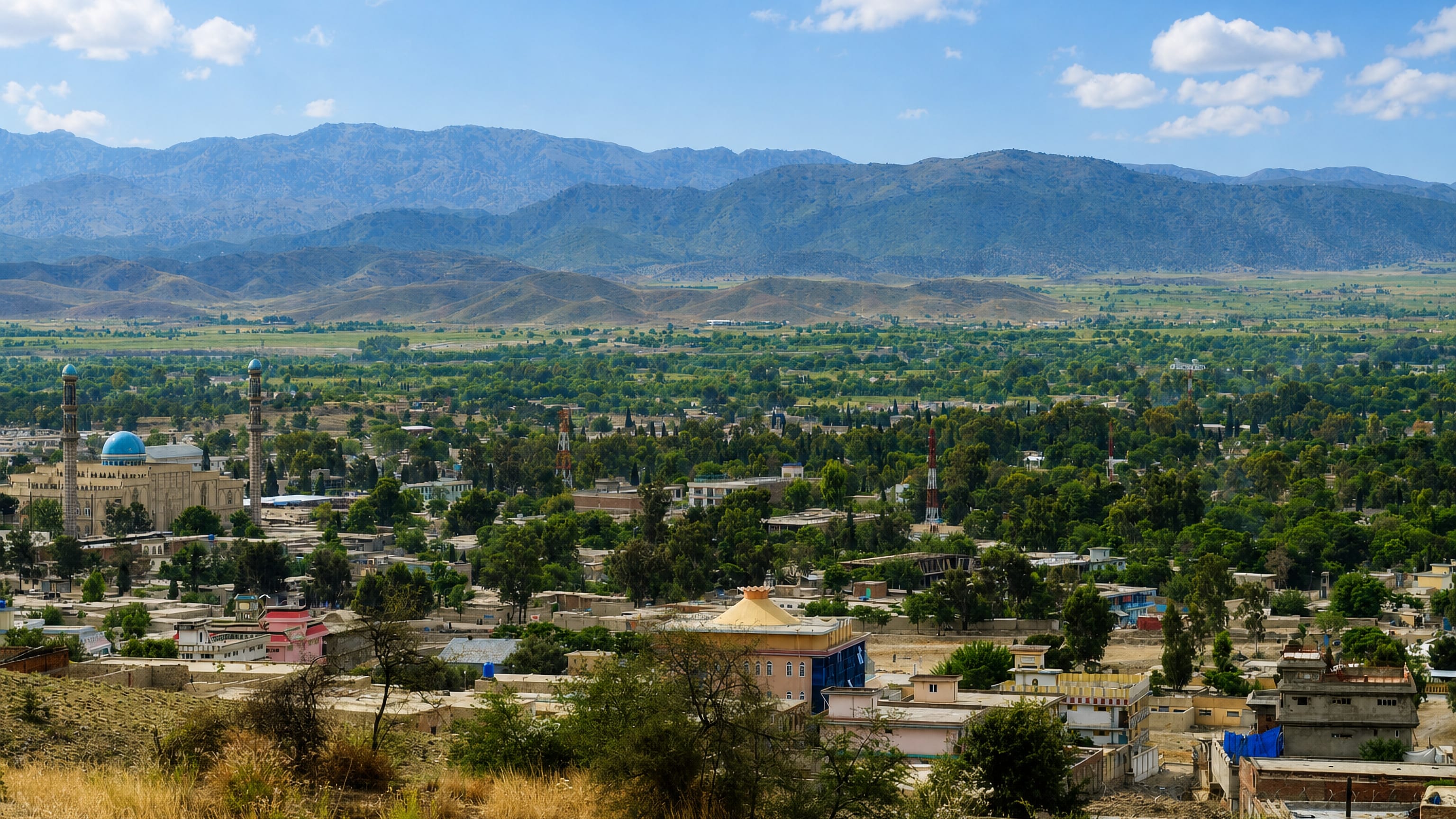 Khost's blue-domed central mosque and low rooftops spread across a green valley beneath the Afghan mountains.
