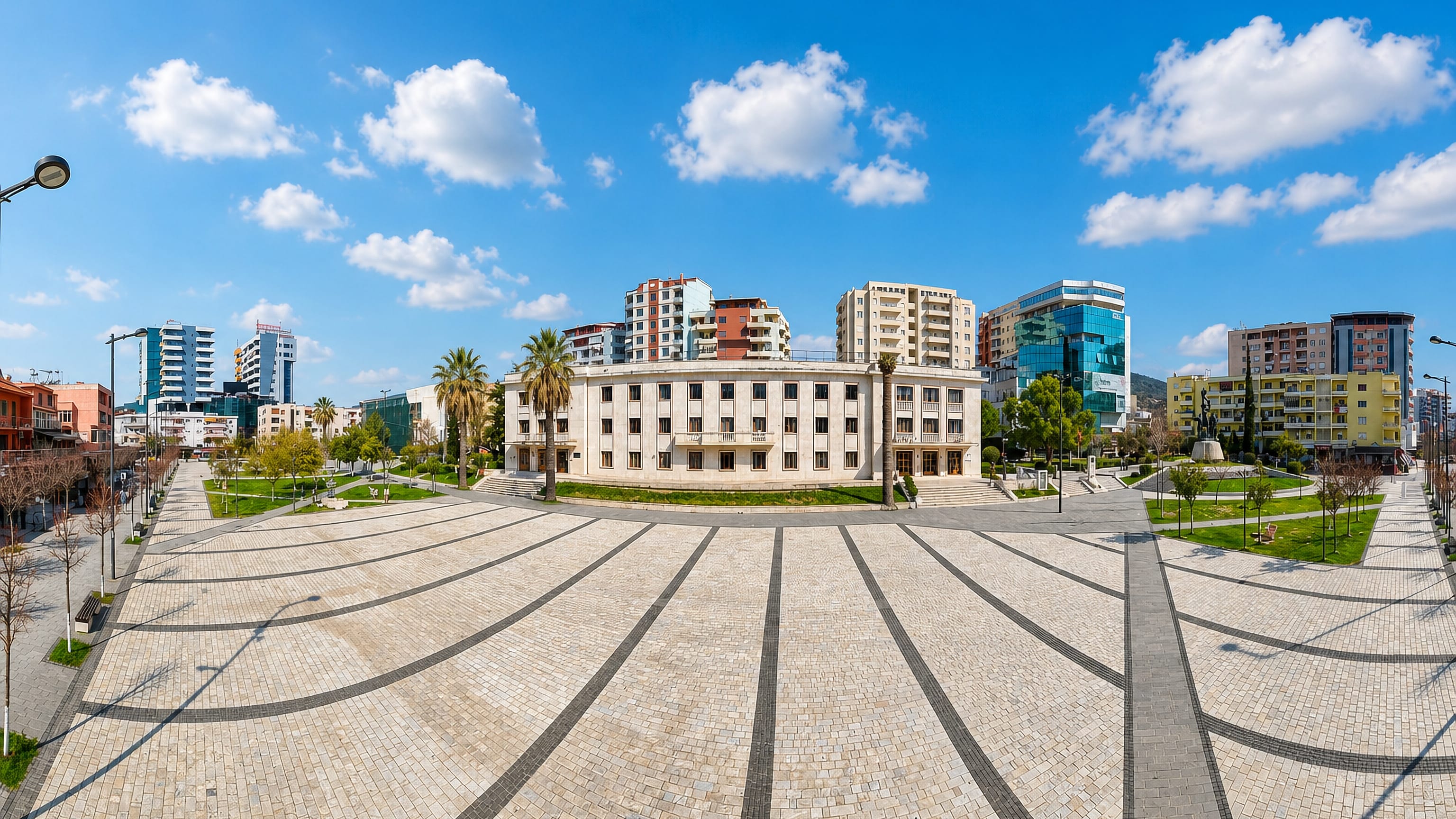 Wide paved central square in Lushnjë, Albania, ringed by apartment blocks and a glass office tower