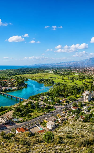Shkodër, Albania, with the turquoise Buna River, a bridge and the Albanian Alps beyond the red-roofed city.