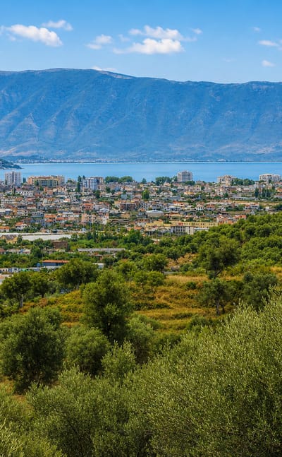 Vlorë, Albania, a coastal city on its bay backed by mountains, with olive groves in the foreground.