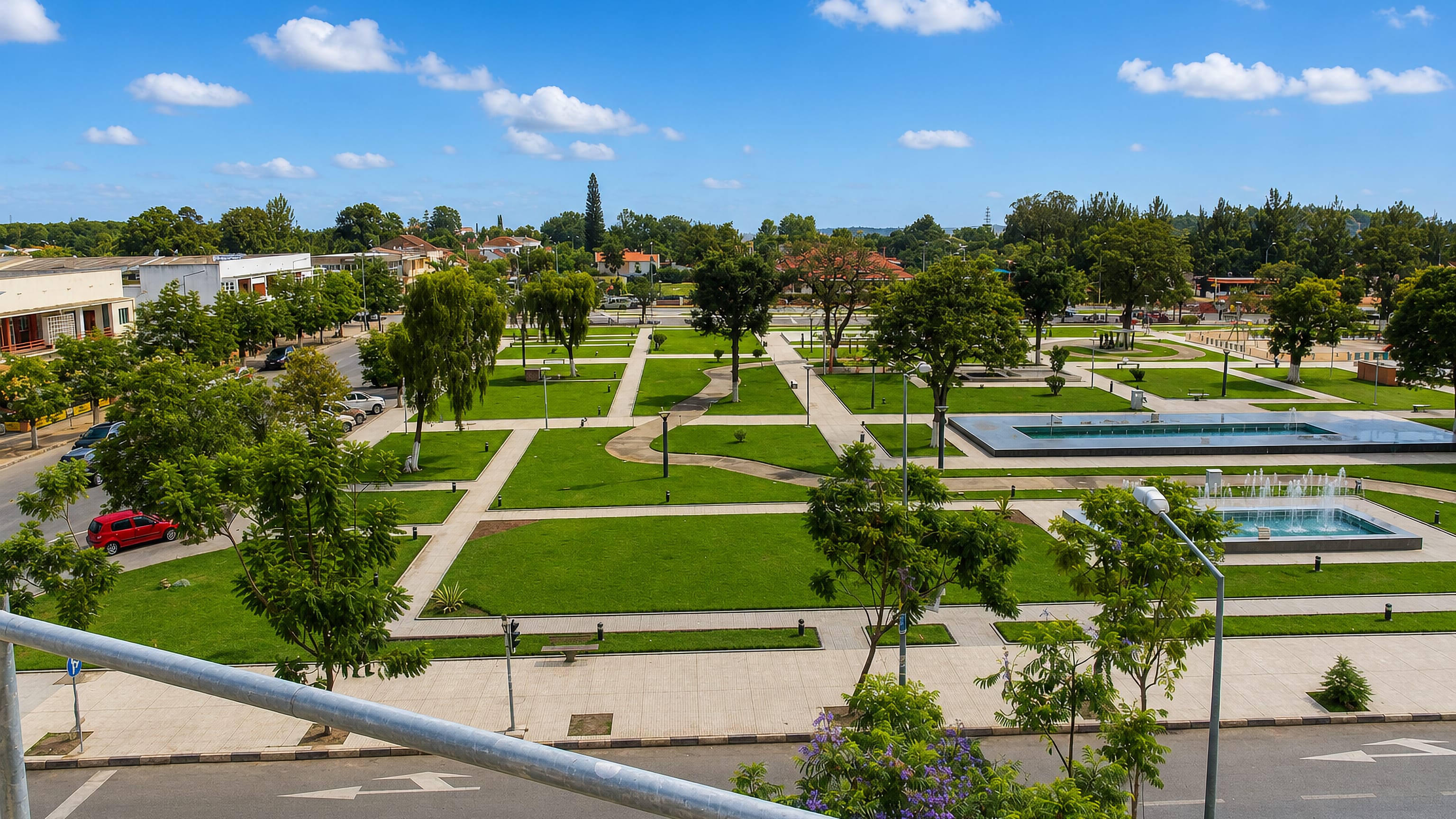 Landscaped central park with fountains and lawns in Huambo, Angola, framed by jacarandas and low buildings.