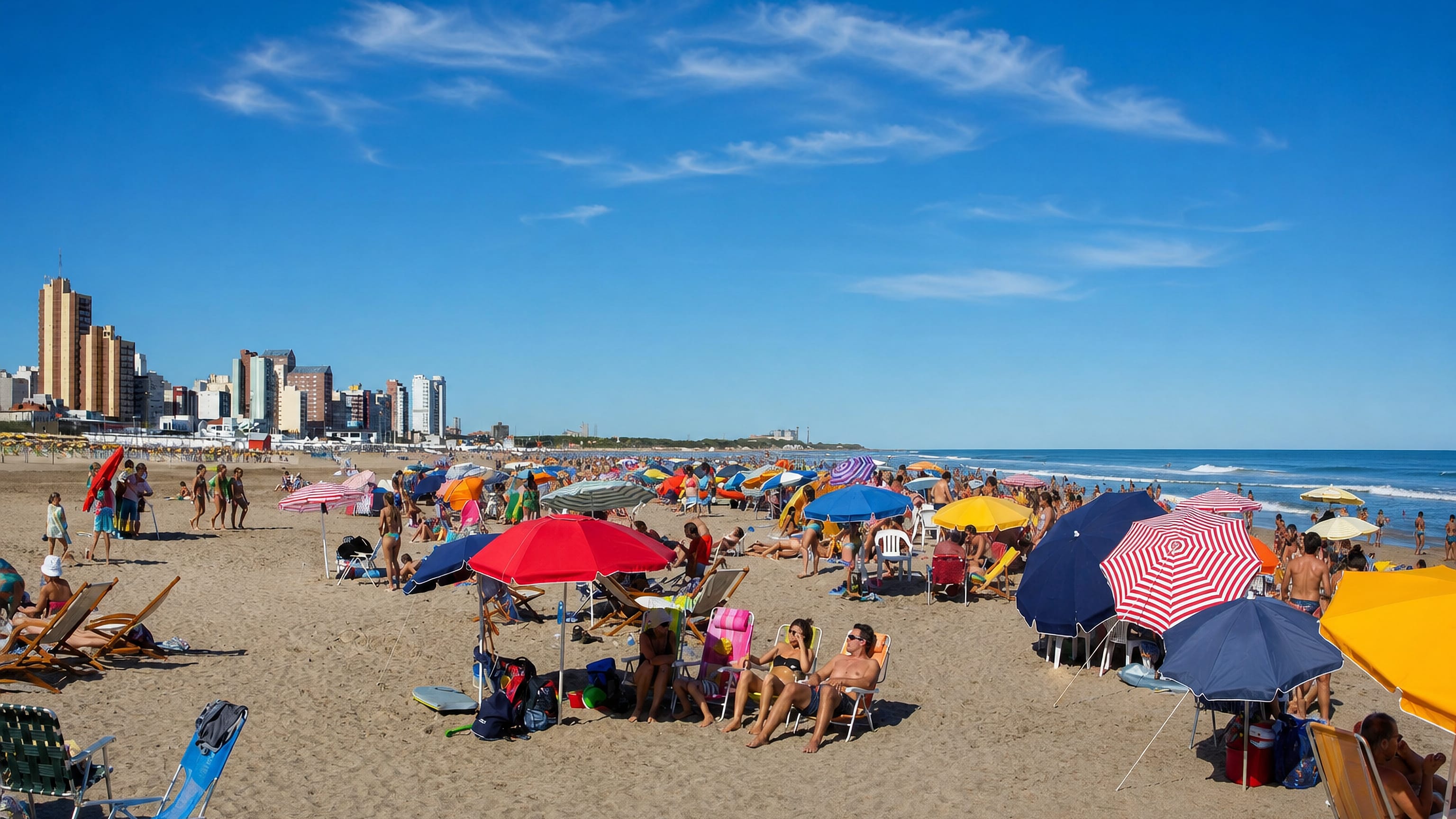 Crowded summer beach at Necochea, Argentina, with sun umbrellas and seafront apartment towers beyond.