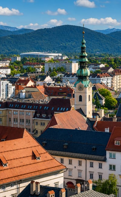 Baroque green-domed church towers and red-tiled rooftops of Klagenfurt am Worthersee with alpine peaks behind.
