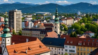 Baroque green-domed church towers and red-tiled rooftops of Klagenfurt am Worthersee with alpine peaks behind.