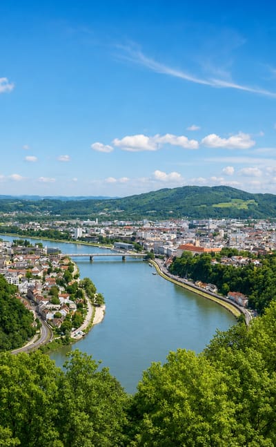 Aerial view of Linz along the Danube, the New Cathedral spire among the rooftops with green hills beyond.