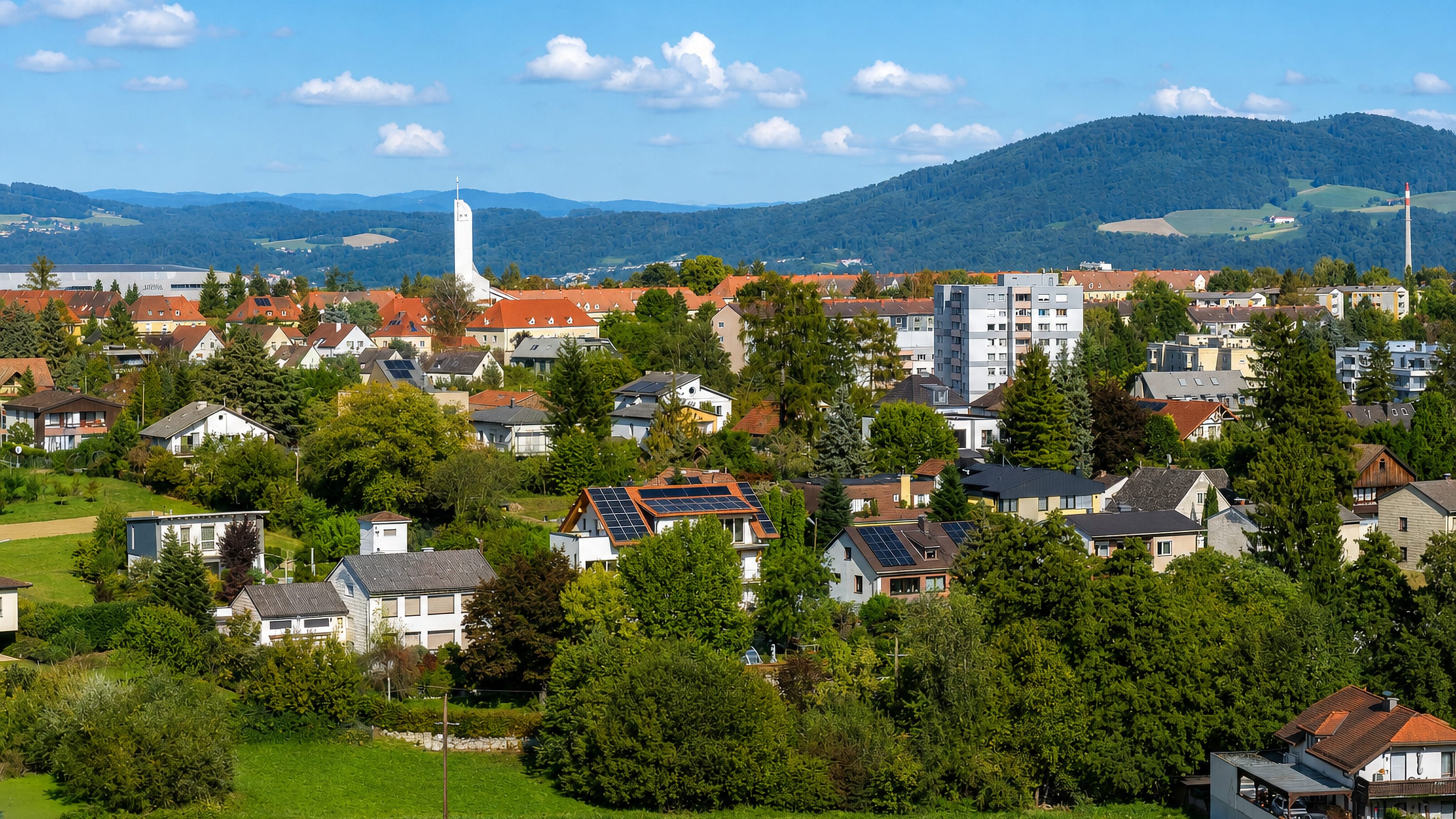 Red-roofed houses and a white church tower below wooded hills in Froschberg, Linz.