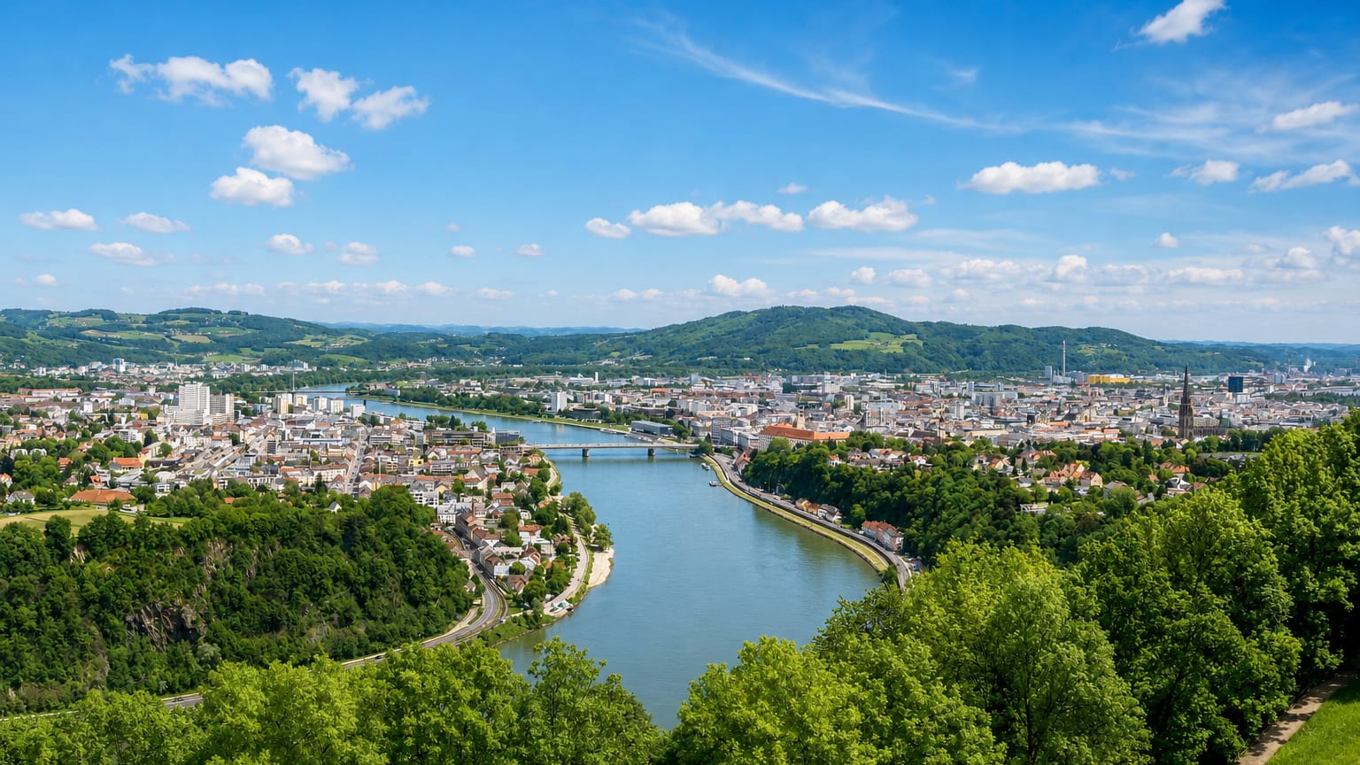 Aerial view of Linz along the Danube, the New Cathedral spire among the rooftops with green hills beyond.