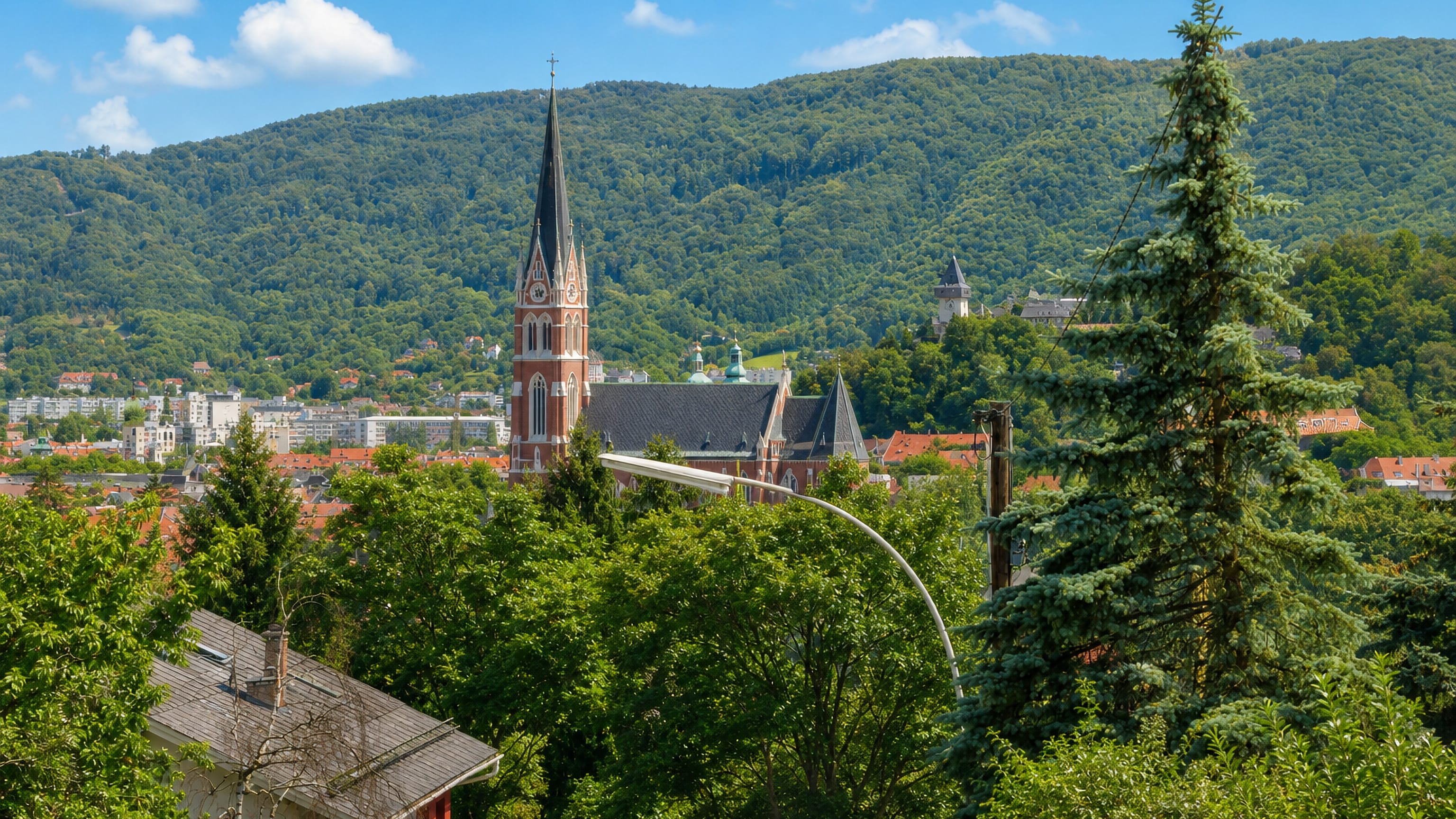 View over treetops from Waltendorf to the Herz-Jesu church spire, Schlossberg clock tower and wooded hills, Graz.