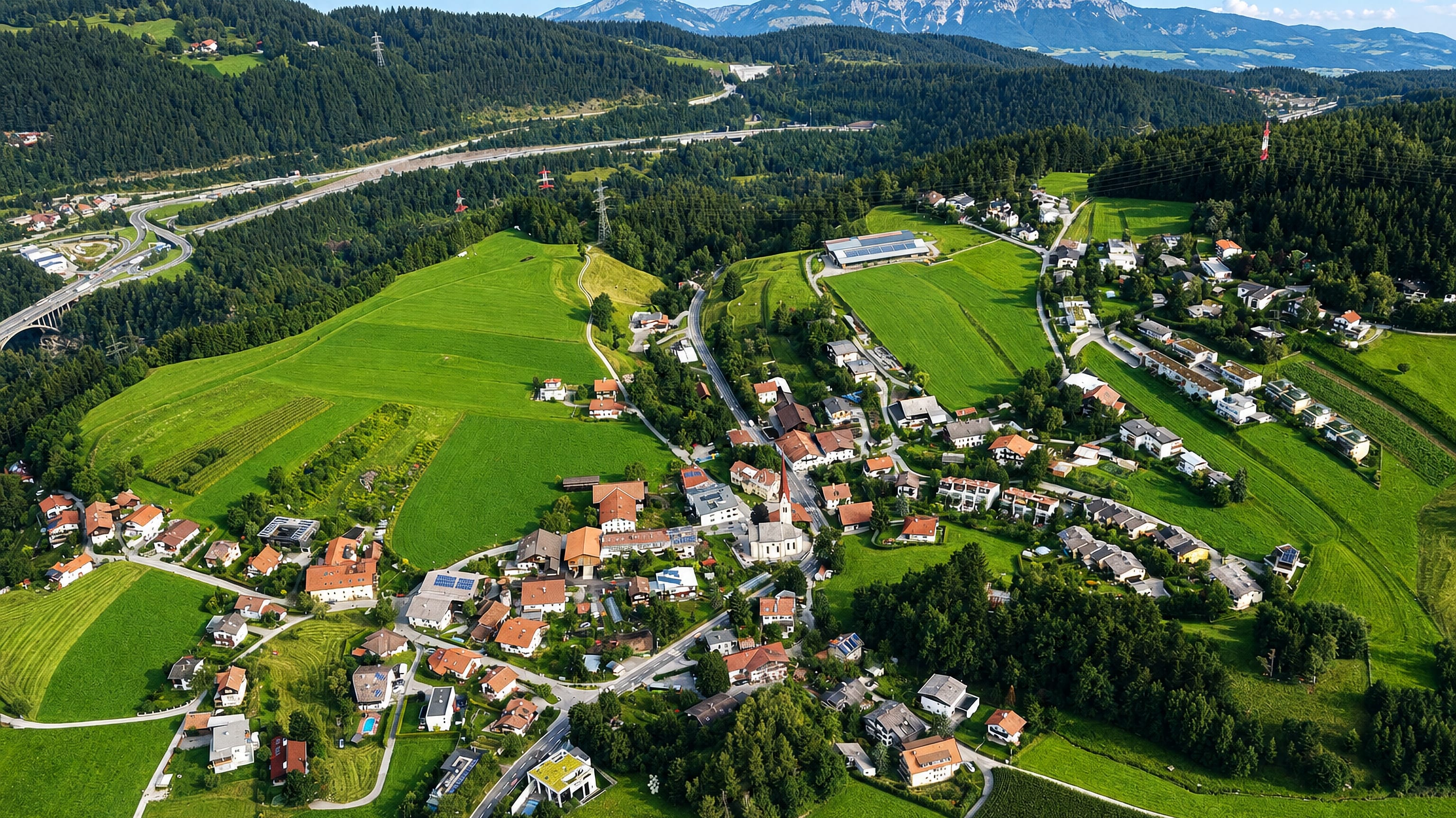 Aerial view of the alpine village of Vill above Innsbruck, its church and meadows ringed by wooded hills.