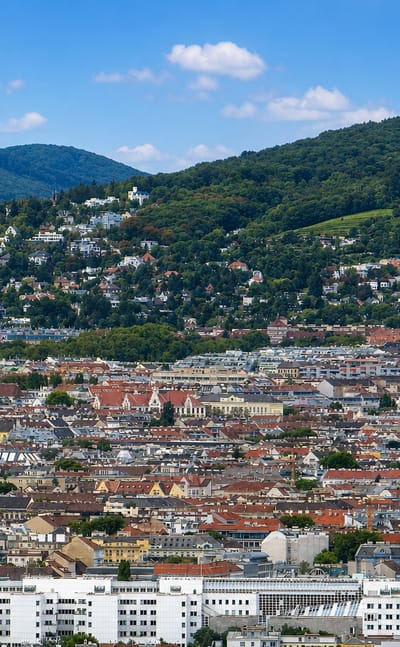 Dense red-tiled Viennese rooftops and church spires backed by the wooded Vienna Woods hills near Alsergrund.
