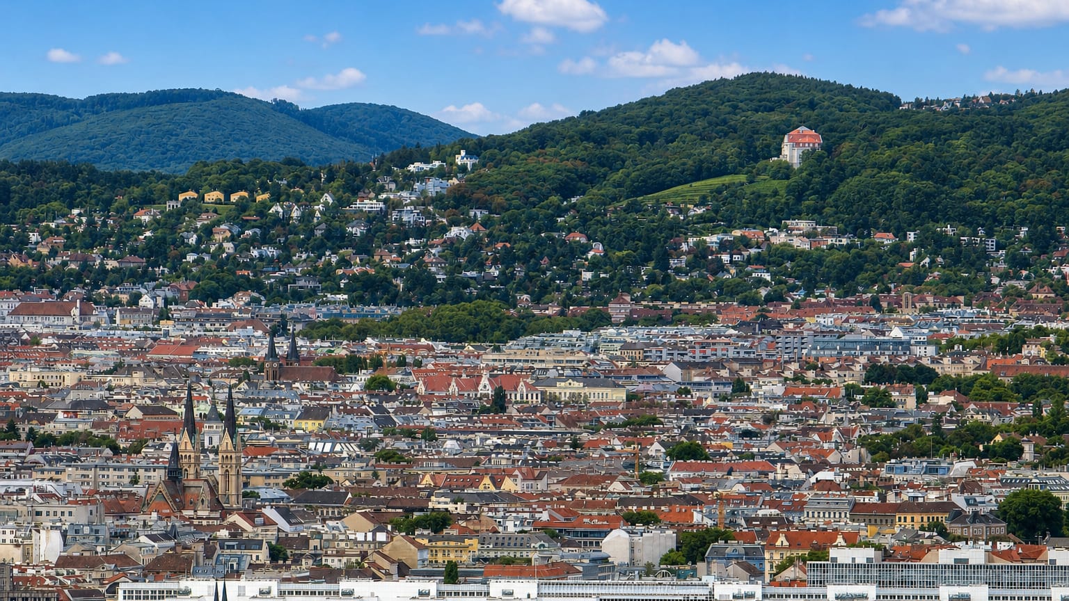 Dense red-tiled Viennese rooftops and church spires backed by the wooded Vienna Woods hills near Alsergrund.