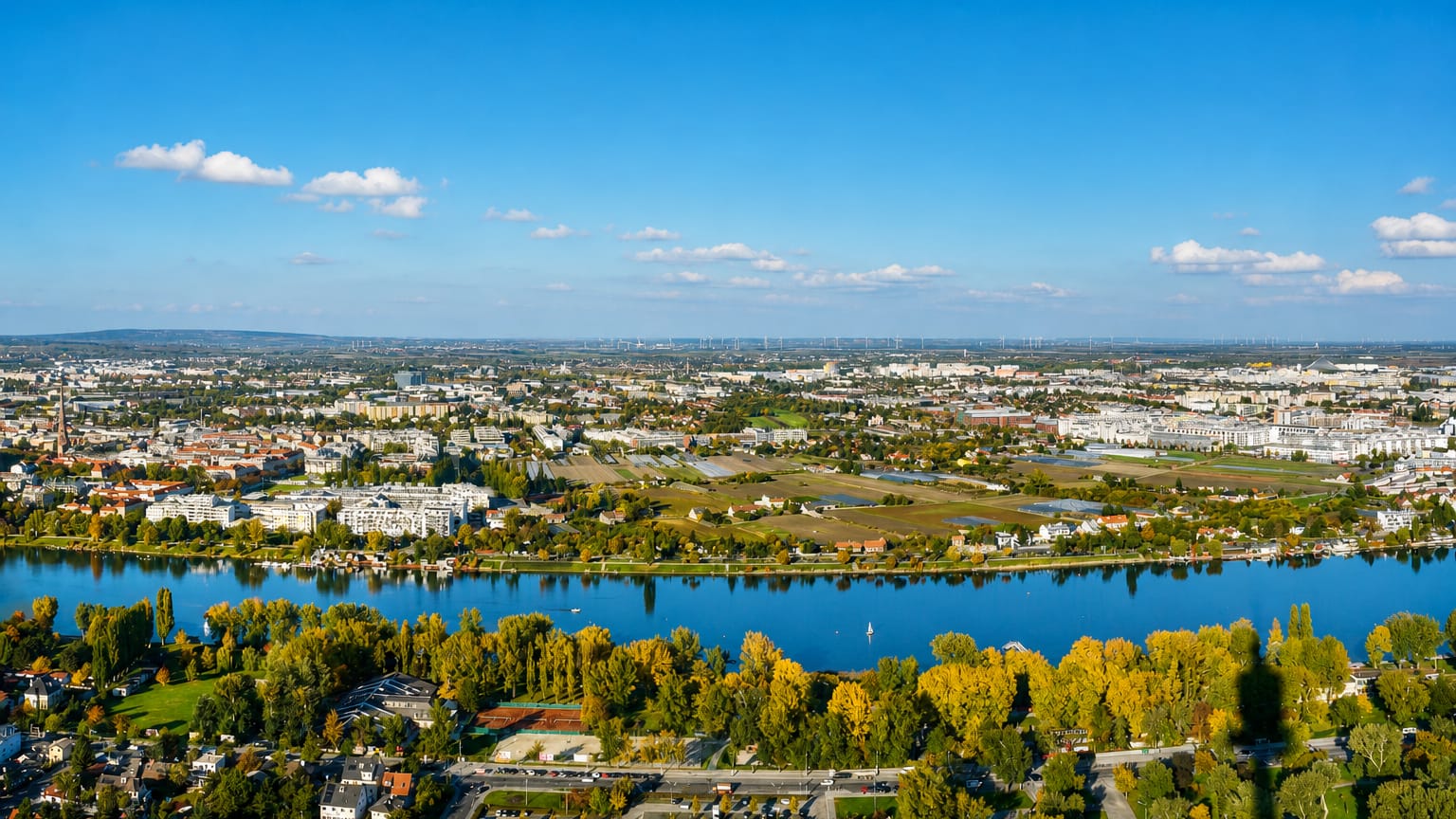 Aerial over the Danube at Floridsdorf, Vienna, with autumn trees, riverside housing and distant wind turbines