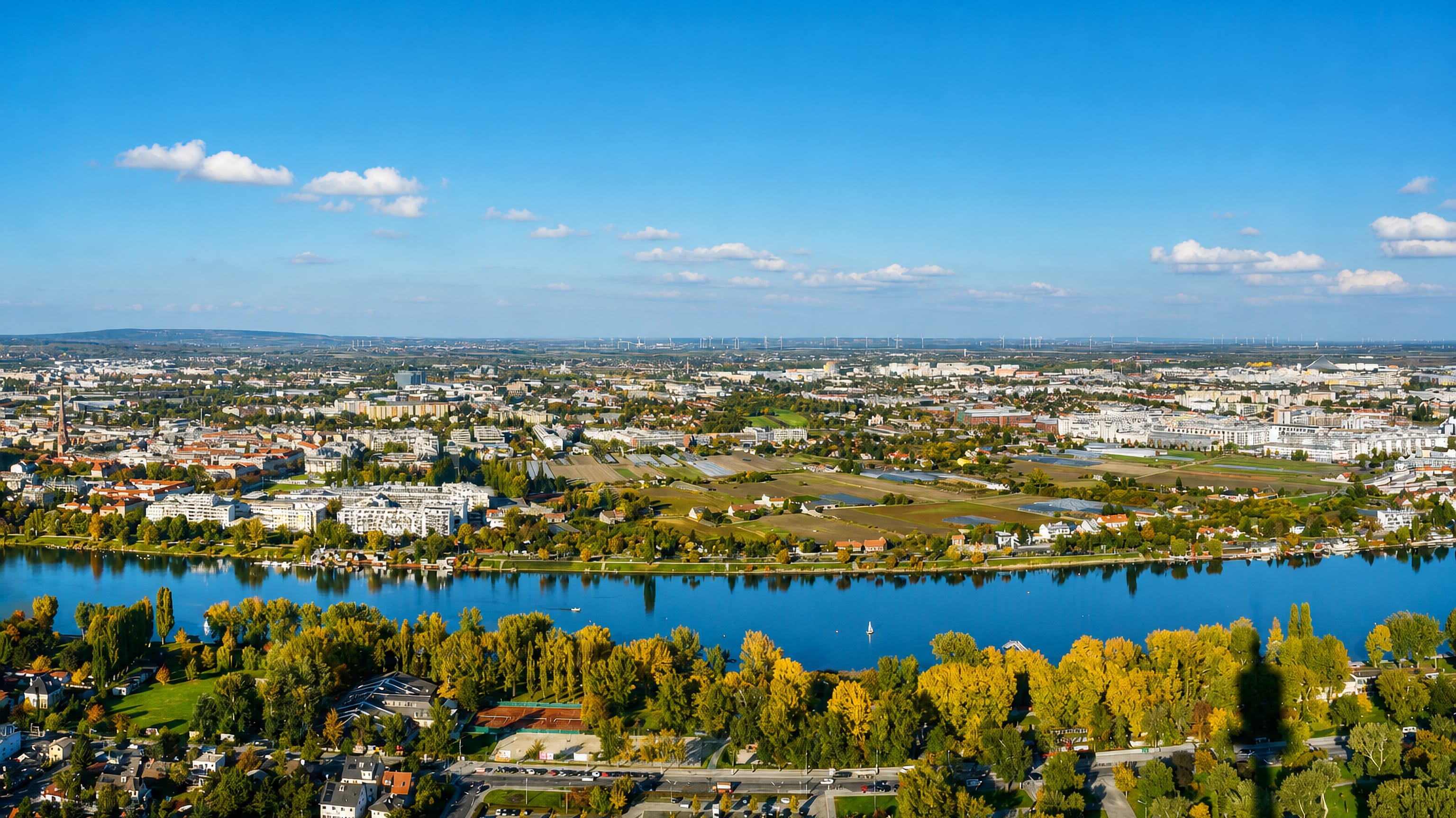 Aerial over the Danube at Floridsdorf, Vienna, with autumn trees, riverside housing and distant wind turbines
