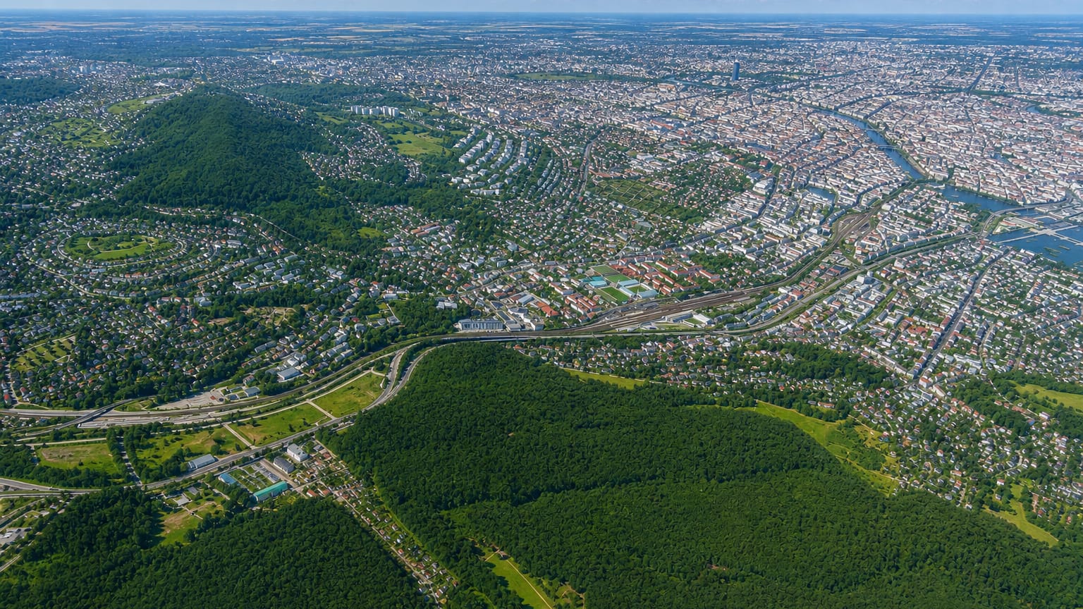 High aerial over western Vienna's Penzing district with the wooded Vienna Woods hills, residential streets and the Danube beyond.