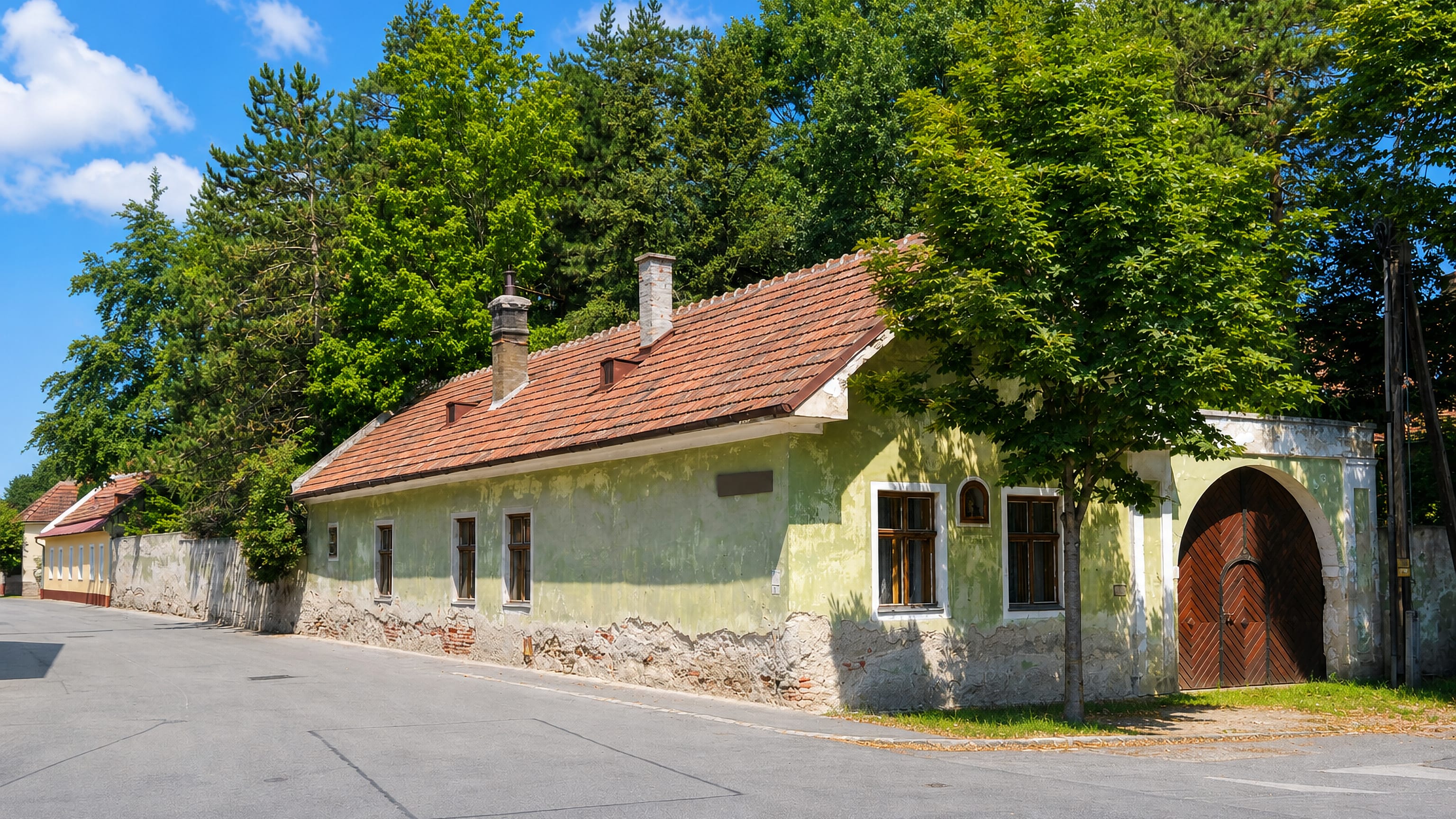 Old single-storey village house with an arched courtyard gate on a leafy lane in Unterlaa, Vienna.