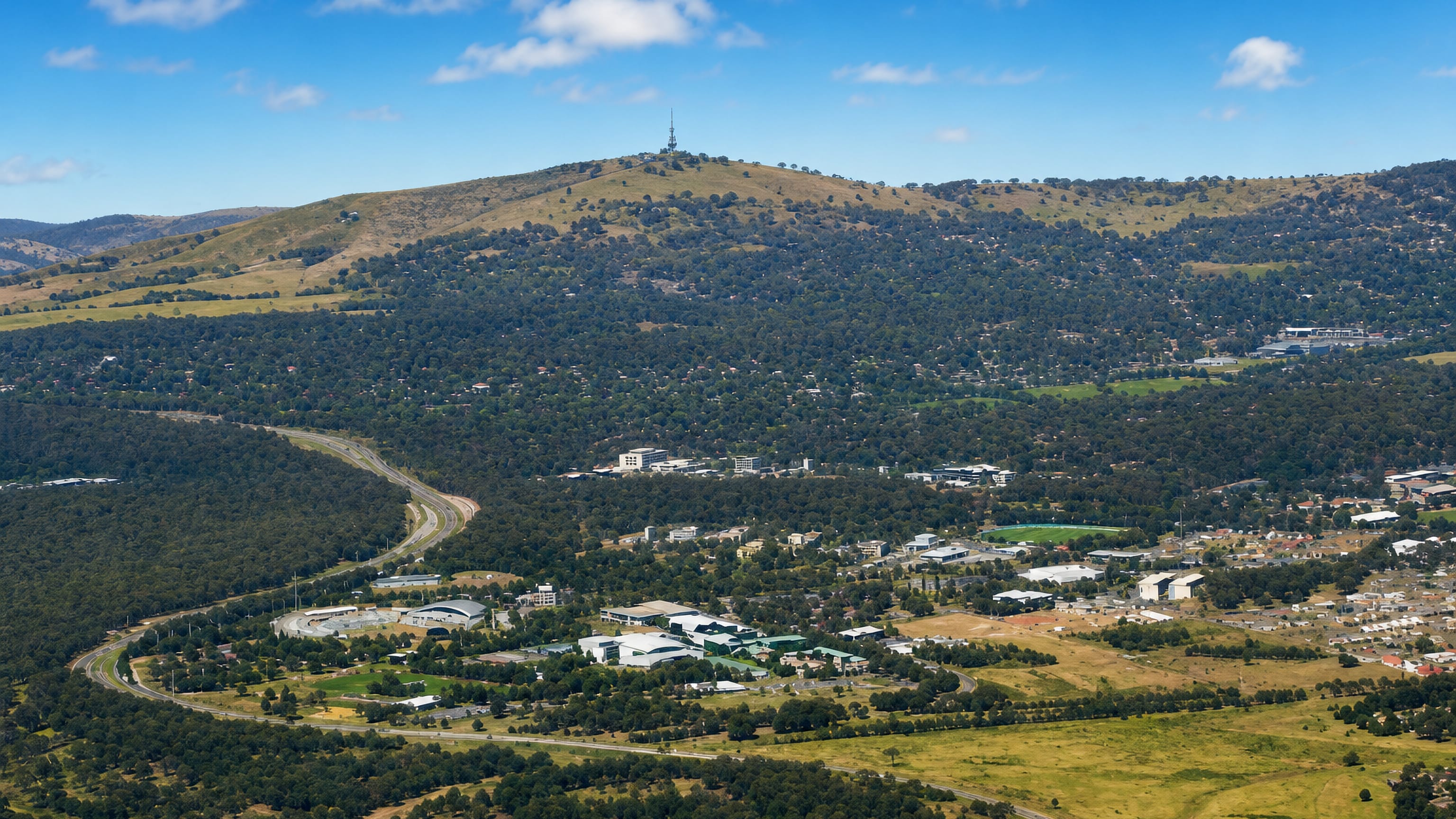 Aerial over Bruce in Canberra, its campus and stadium set in bushland below Black Mountain's Telstra Tower.