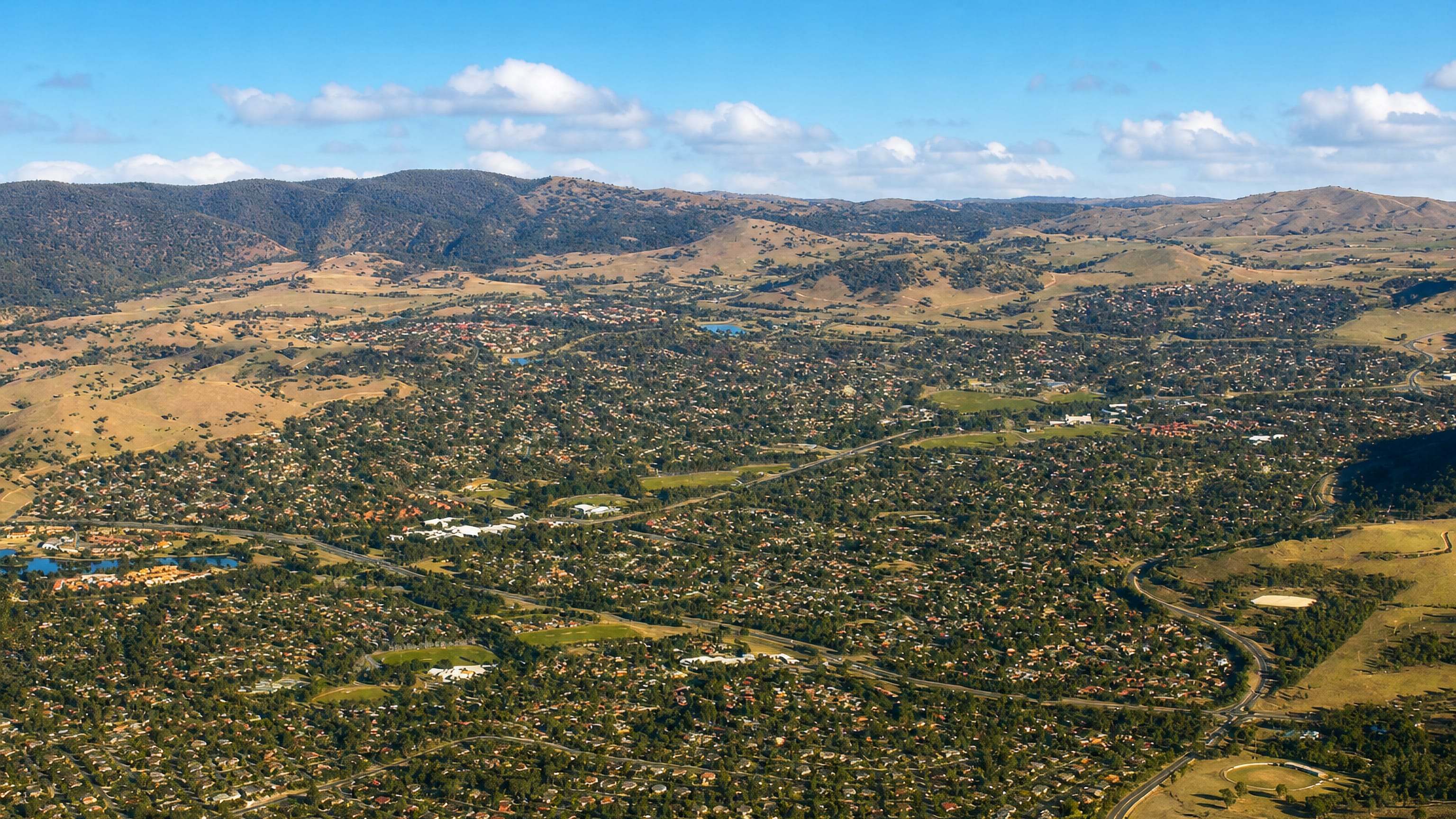 Aerial view of Kambah's tree-covered suburban streets in Canberra's Tuggeranong valley below dry hills.