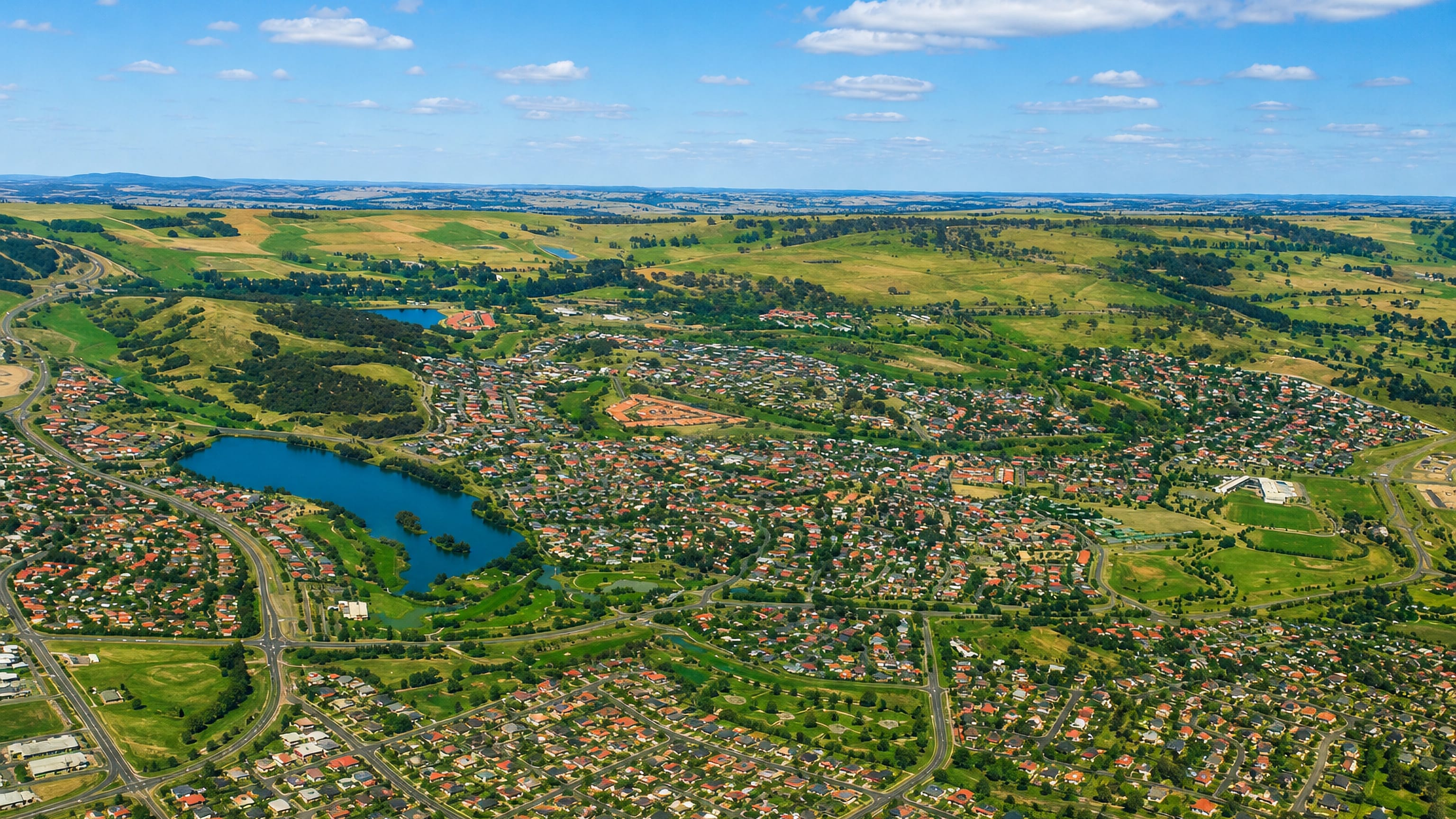 Aerial view of Nicholls, Canberra, with tiled-roof houses around a lake and open grassland hills beyond.