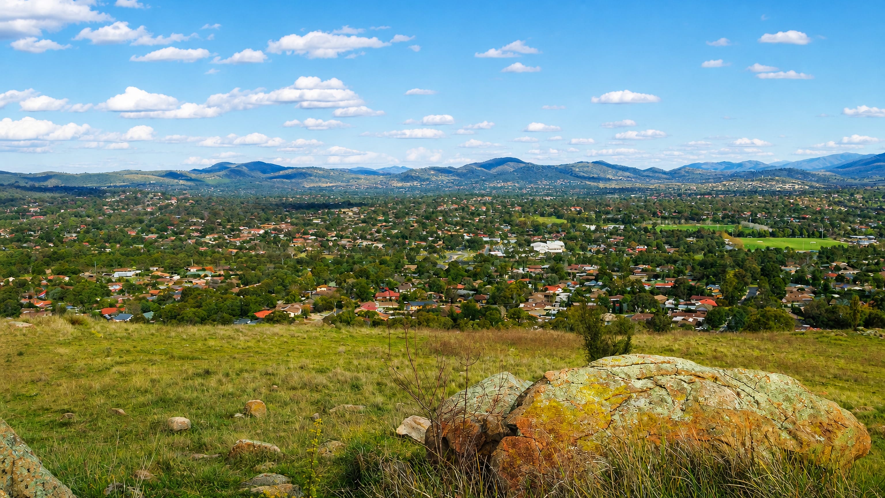 View from a grassy, rock-strewn hilltop over the suburban streets of Wanniassa, Canberra, to distant ranges.