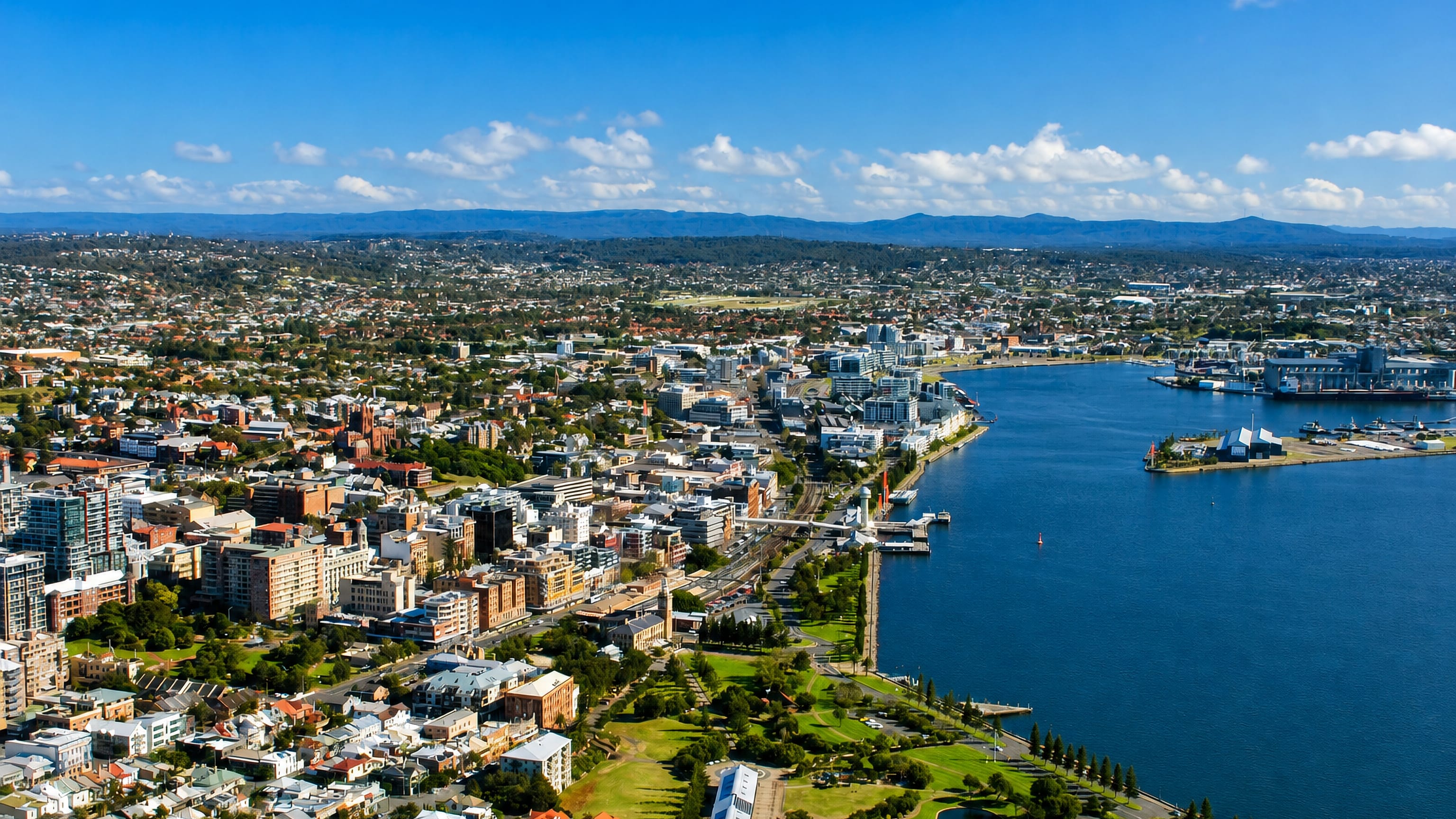 Aerial view of Newcastle, NSW, with its harbour channel, foreshore parkland and CBD backed by distant ranges.