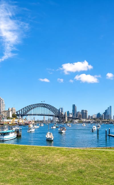 Sydney Harbour Bridge and the city skyline across the harbour, with moored boats and a grassy foreshore in Sydney.