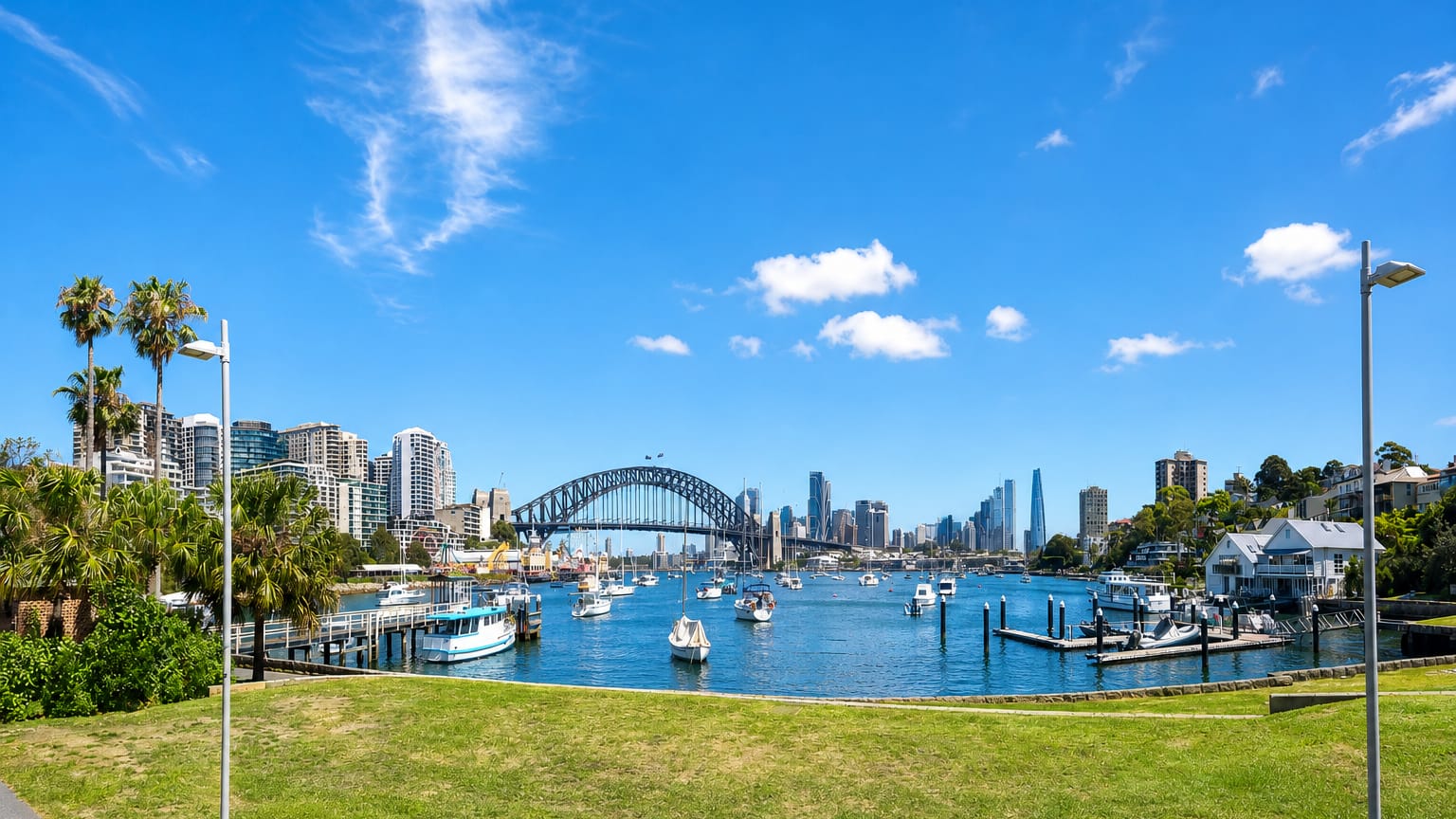 Sydney Harbour Bridge and the city skyline across the harbour, with moored boats and a grassy foreshore in Sydney.