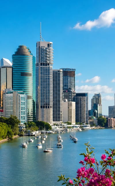 Brisbane central business district skyline rising above the Brisbane River, with moored sailboats and bougainvillea in the foreground