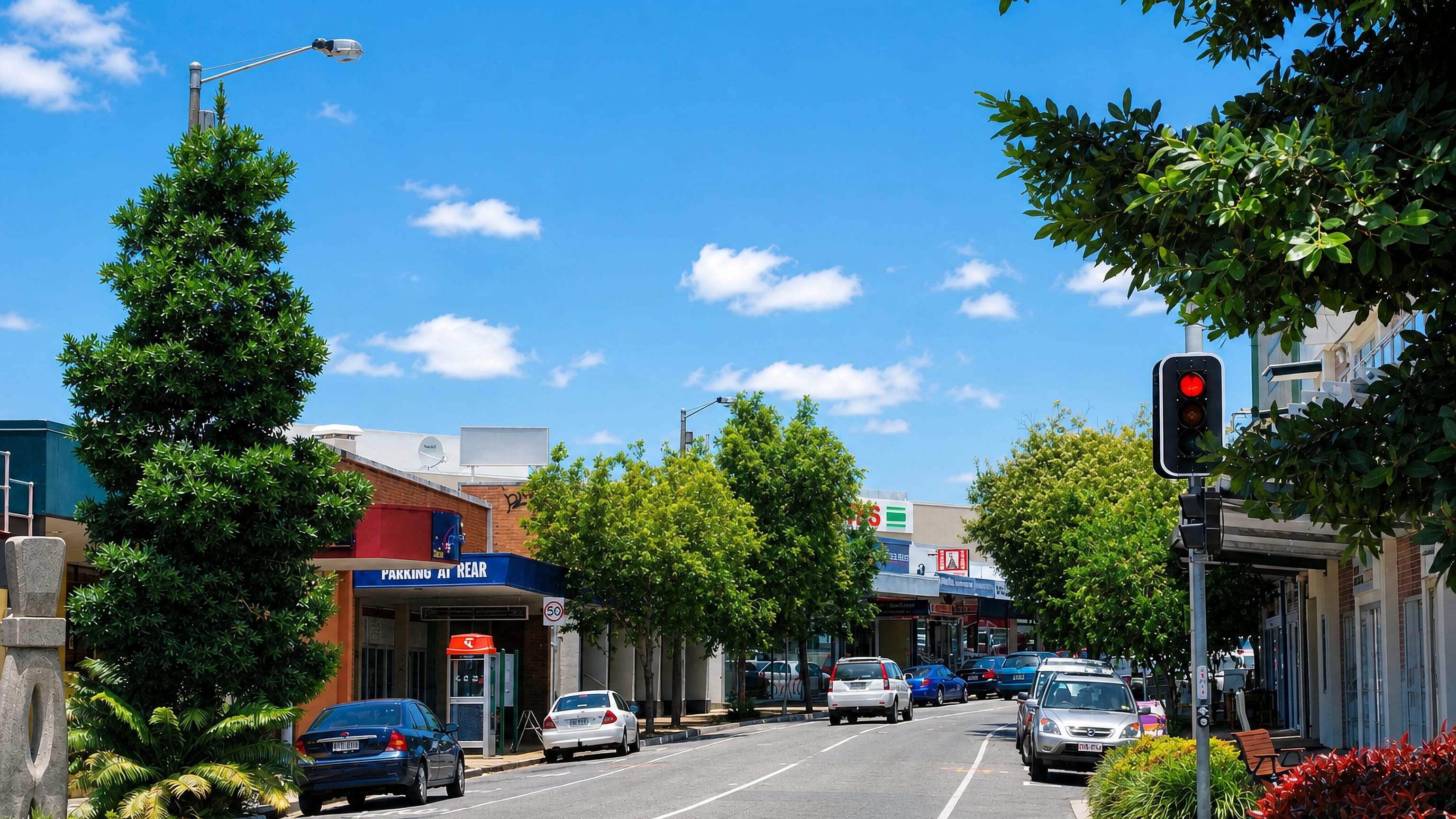 Tree-lined shopping street with shopfronts and parked cars in the Brisbane suburb of Nundah.