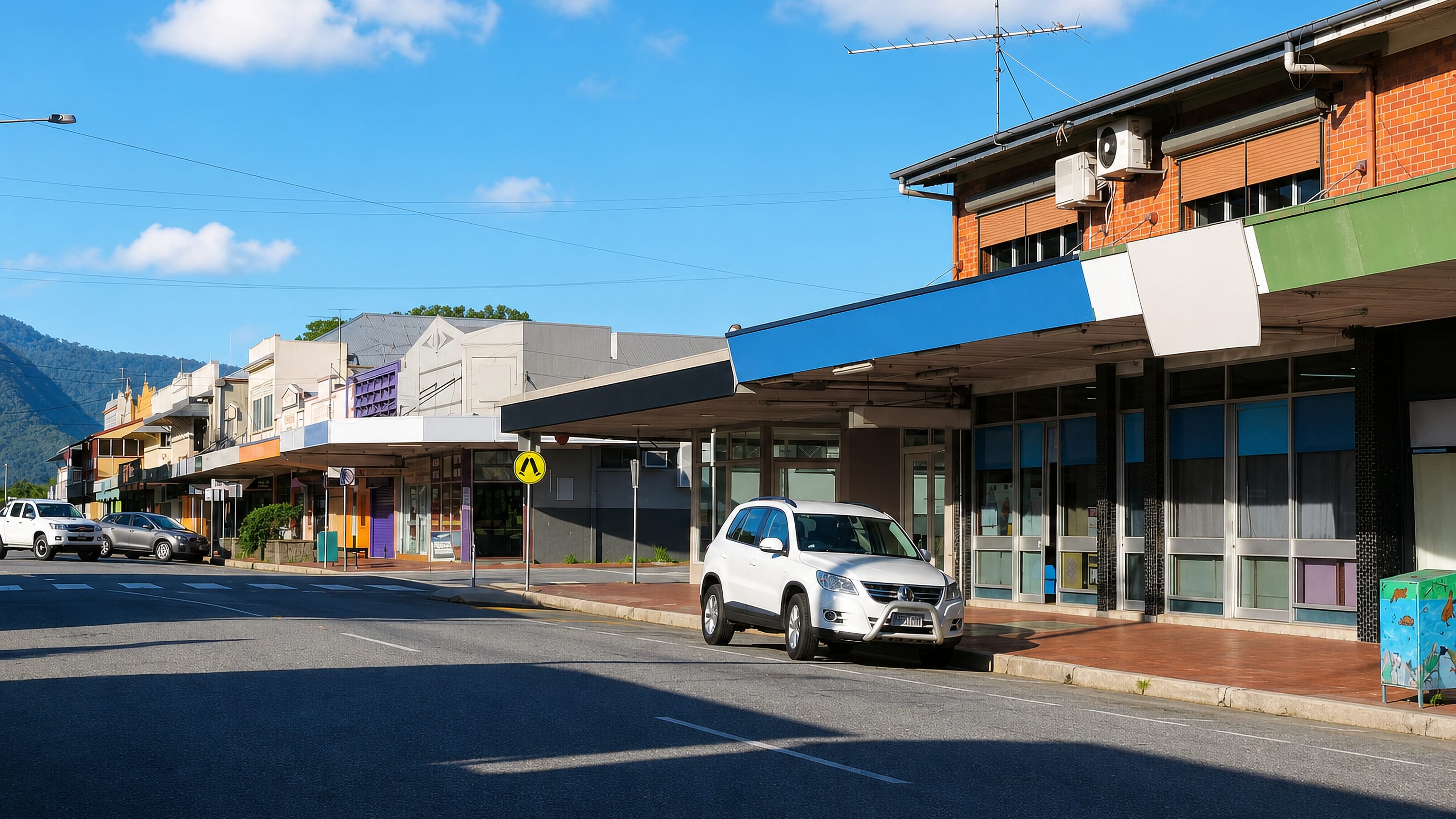 Awning-shaded shopfronts along the main street of Gordonvale, Queensland, with rainforest hills behind.