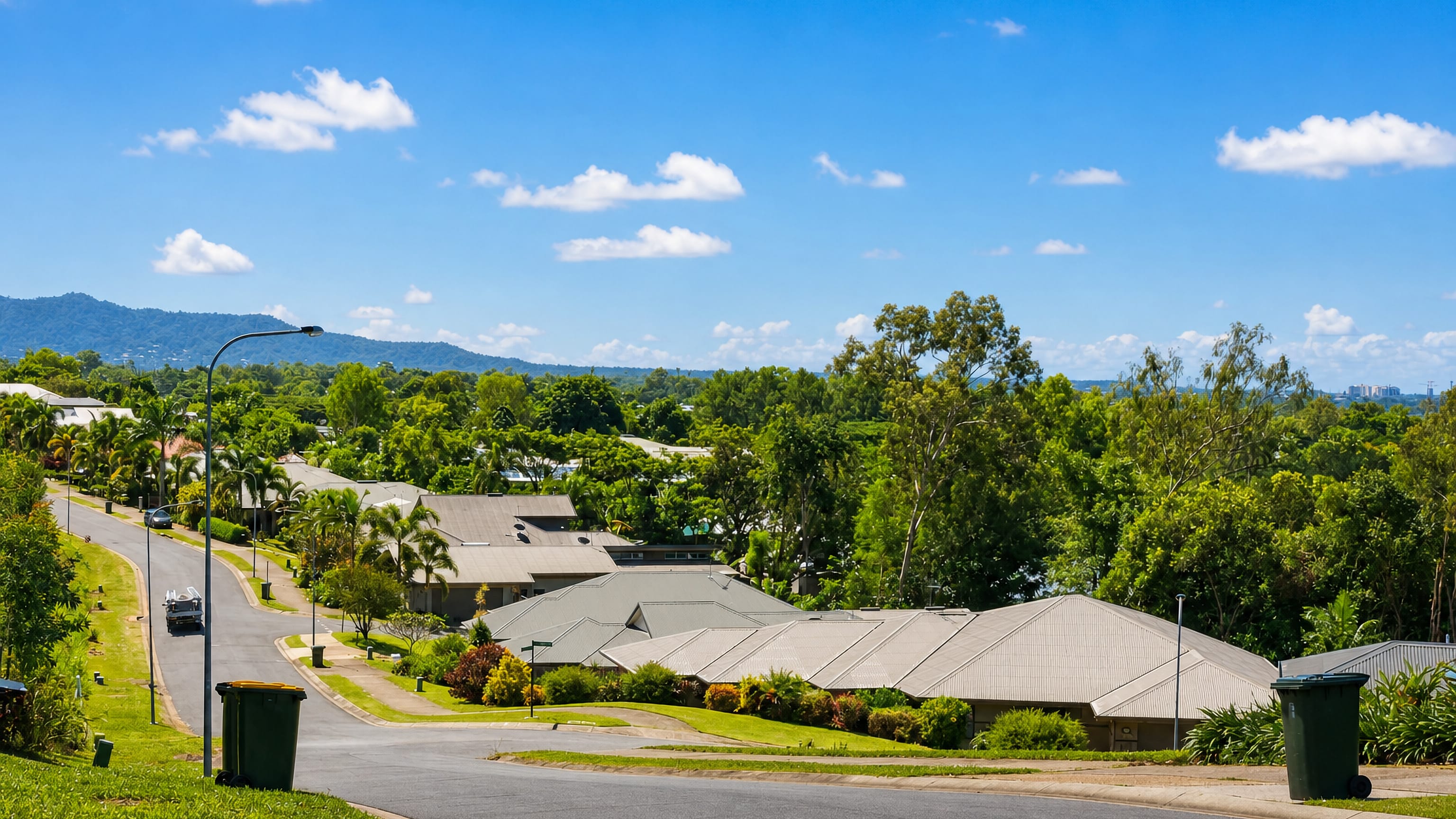 Sloping suburban street with metal-roofed houses, palms and distant ranges at Woree, Cairns, Australia.