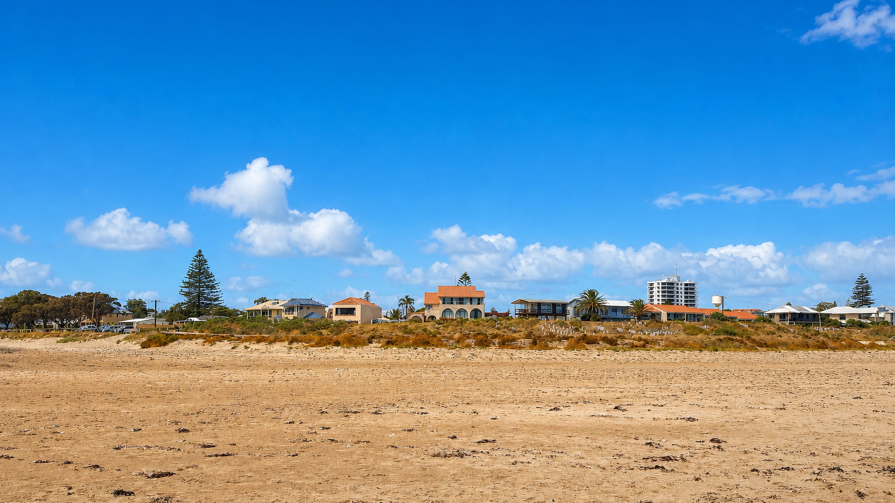 Wide sandy beach at Halls Head, Mandurah, backed by dunes, low coastal homes and a white apartment tower.