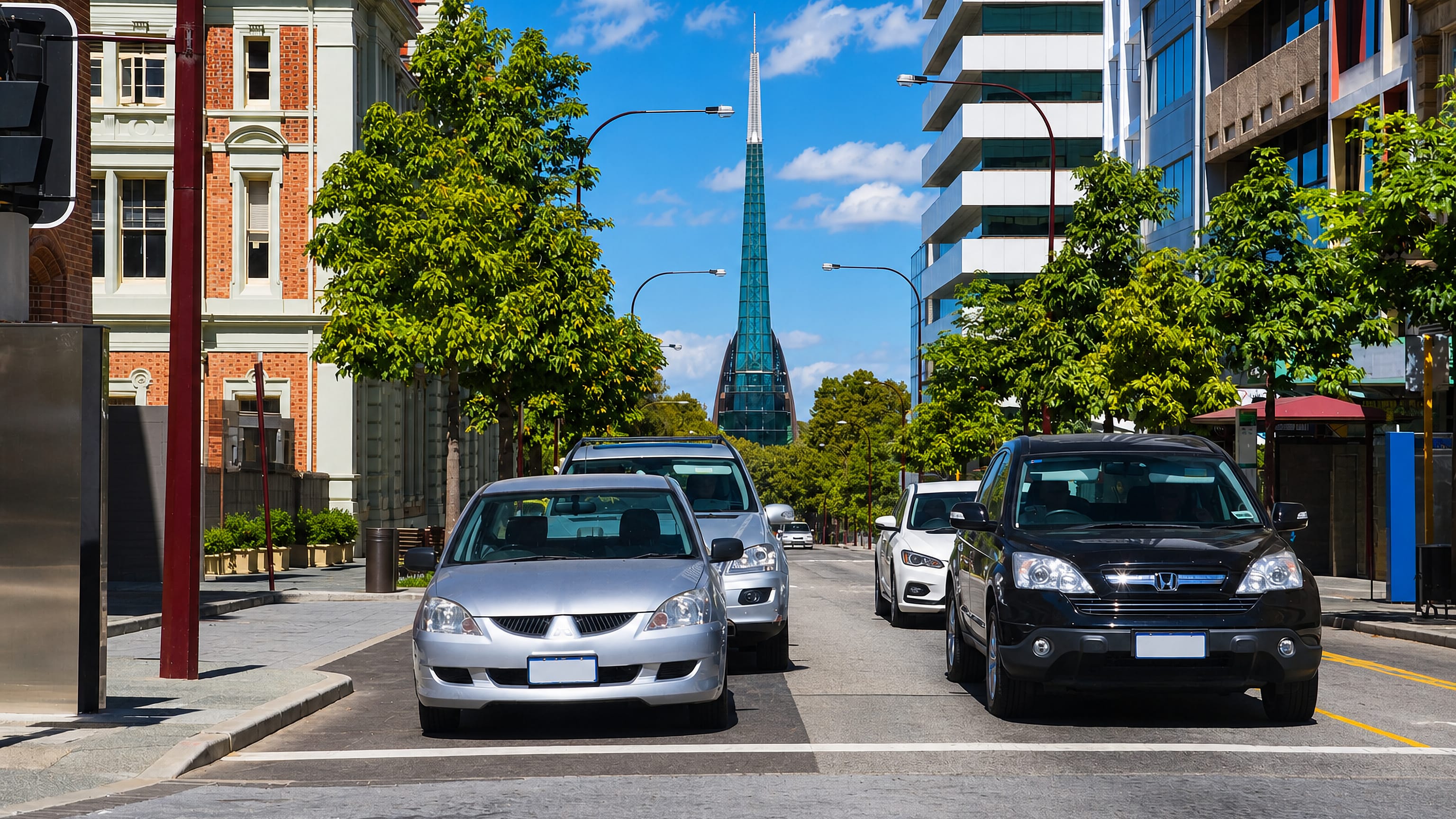Perth city centre street framing the Swan Bells tower spire, with parked cars and shade trees along the kerb.