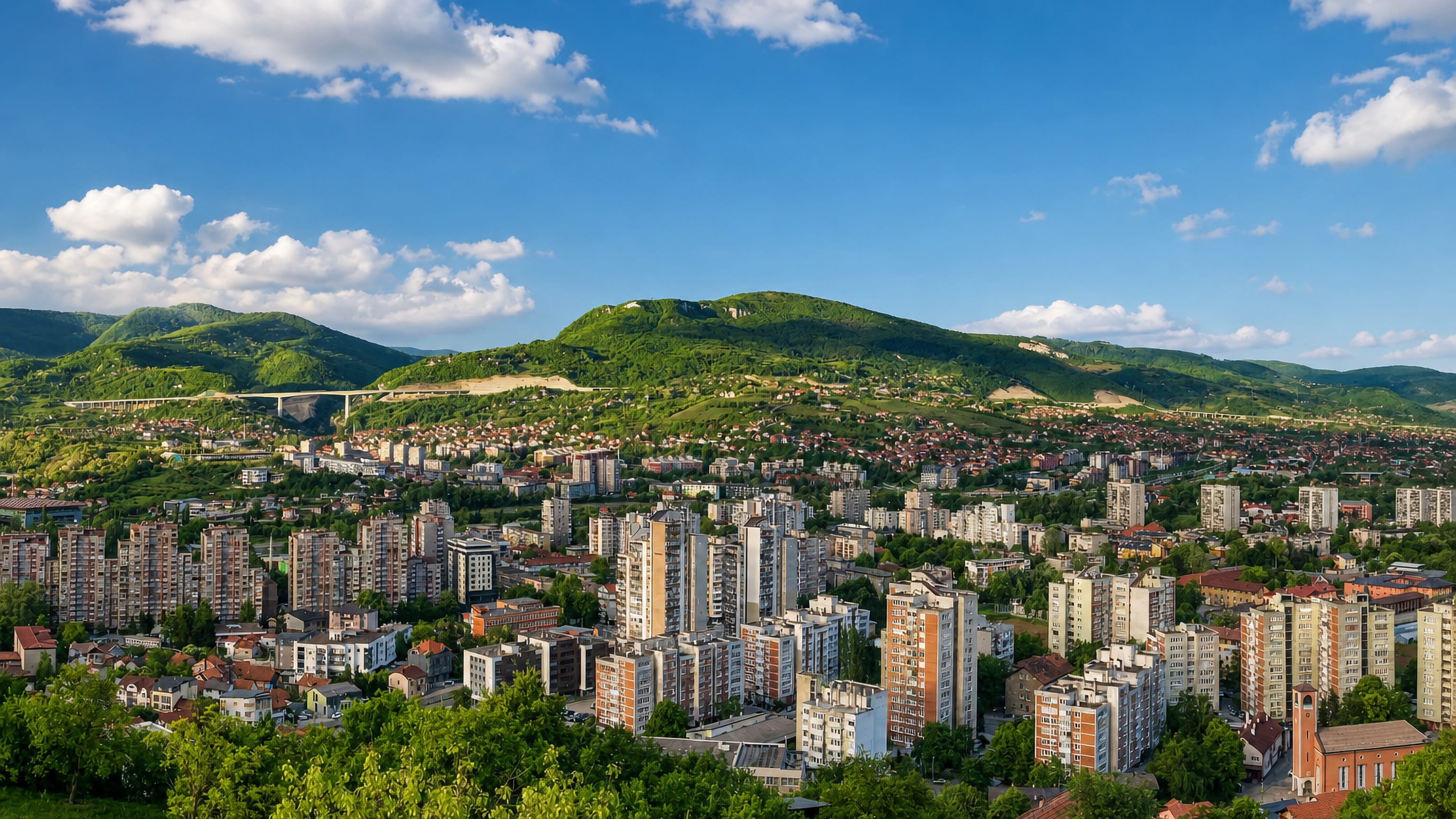 Zenica, Bosnia and Herzegovina, seen across its valley of high-rise blocks, with green hills and a motorway viaduct.