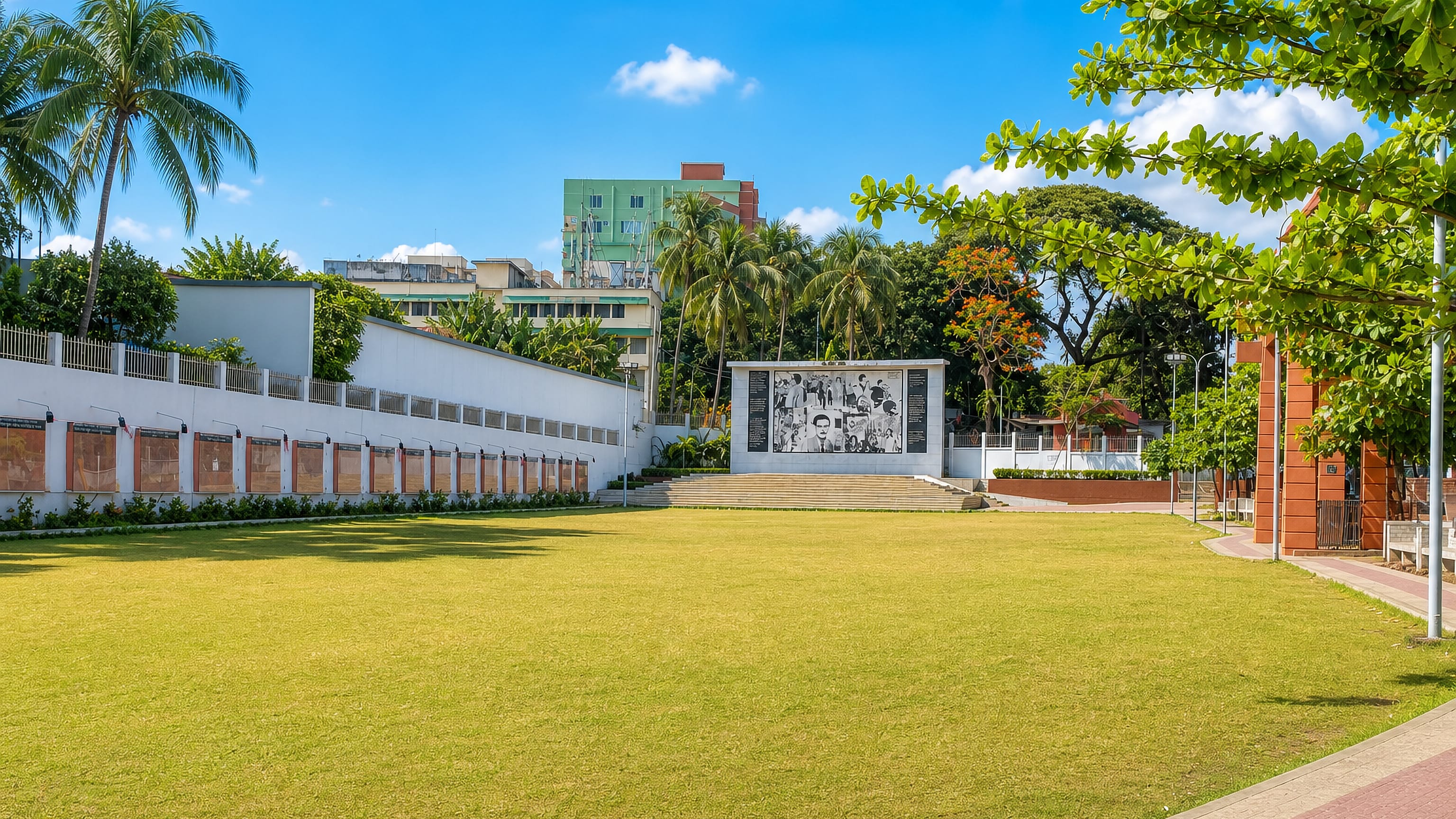 Memorial mural and colonnade beside a lawn at Laldighi in Chattogram, framed by palm trees.