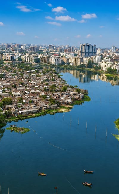 Aerial view of Dhaka with a lake dividing dense informal riverside settlements from mid-rise city towers under a blue sky.