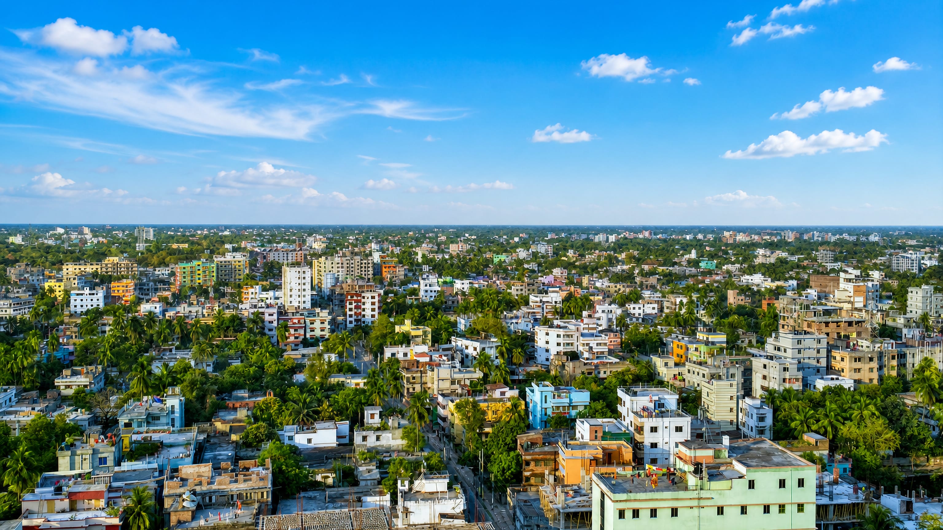 Rajshahi, Bangladesh, a low-rise cityscape of colourful concrete houses among palms under a bright blue sky.
