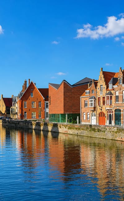 A Bruges canal lined with stepped-gable brick houses reflected in still water under a blue sky.