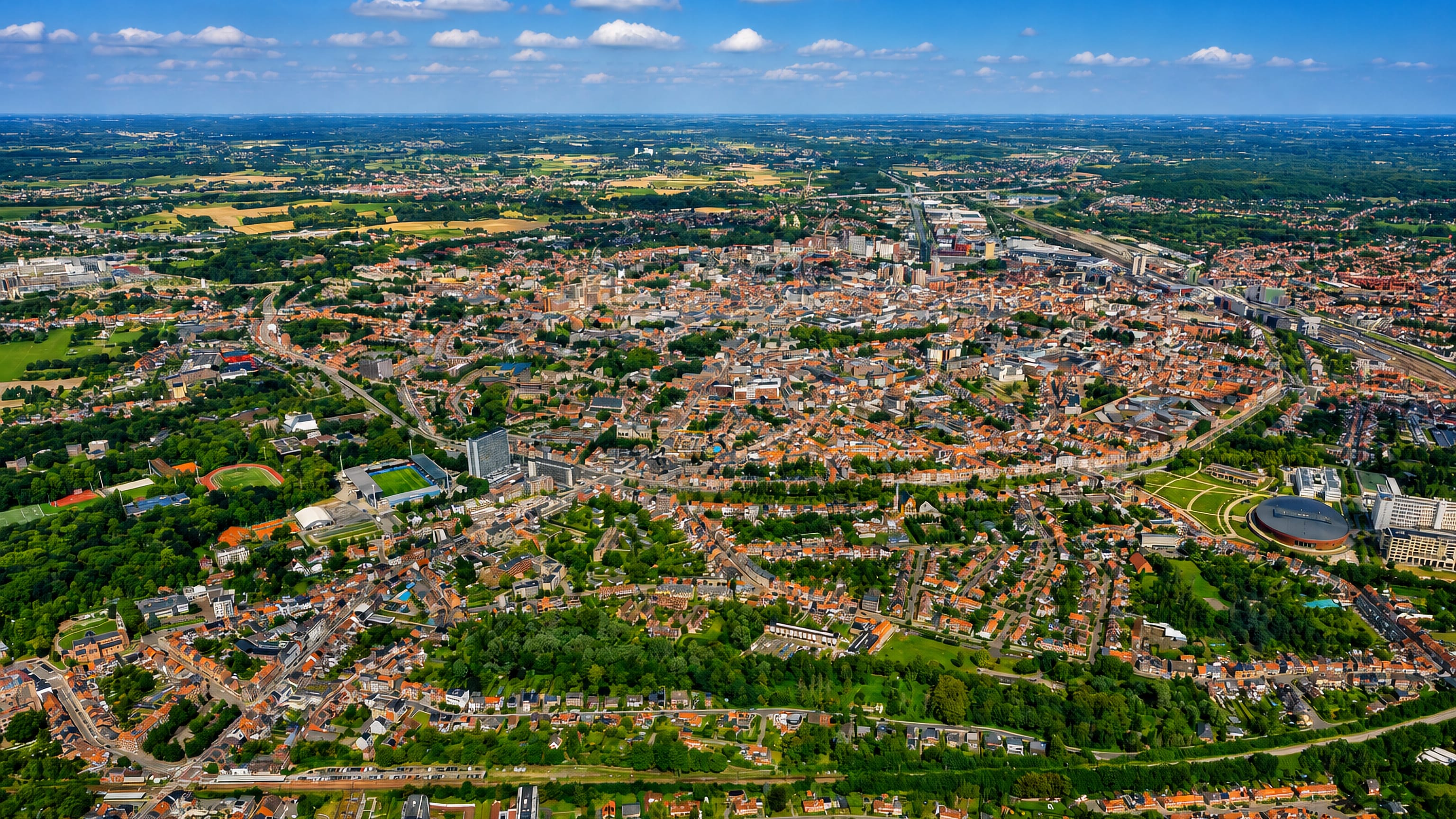 Aerial view over Leuven's dense red-roofed centre, ring road and surrounding Flemish Brabant farmland.