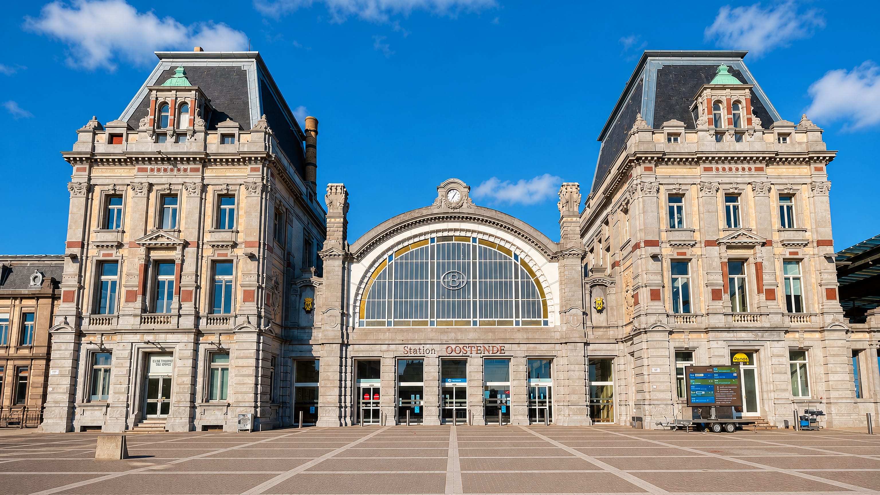 Ostend's neo-Renaissance Station Oostende and its brick-paved forecourt under a clear blue sky.