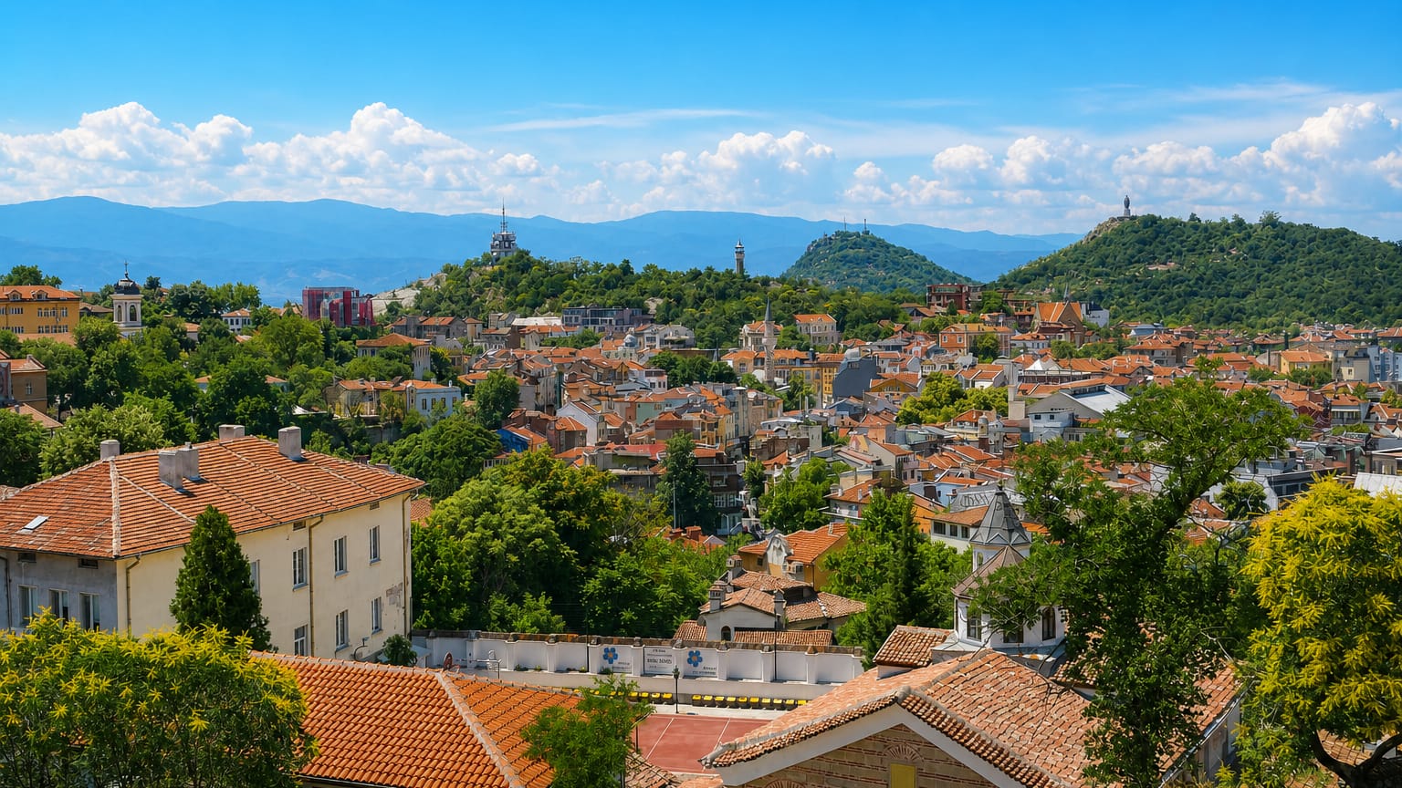 Panorama over Plovdiv's red-tiled roofs and wooded hills topped by hilltop monuments.