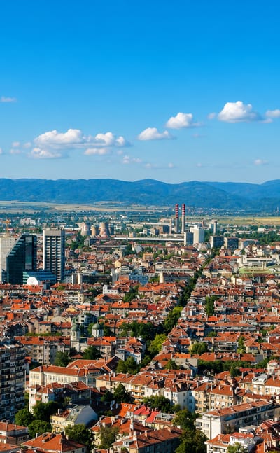 Aerial view of Sofia's dense red-roofed streets stretching toward the Balkan mountains under a clear sky.