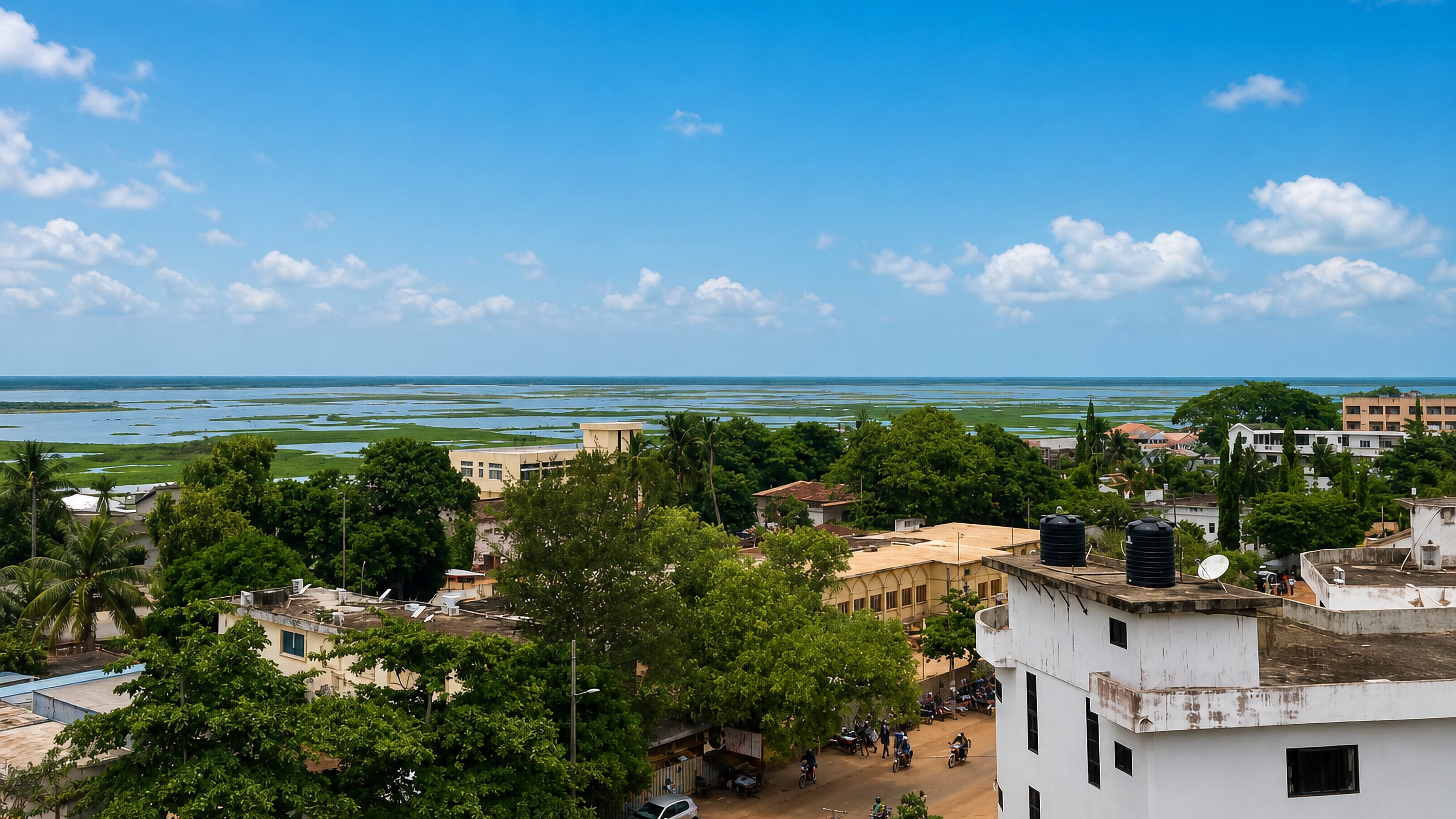 Rooftop view over Porto-Novo's low-rise centre toward the green Nokoué lagoon, with motorbikes on a sandy street.