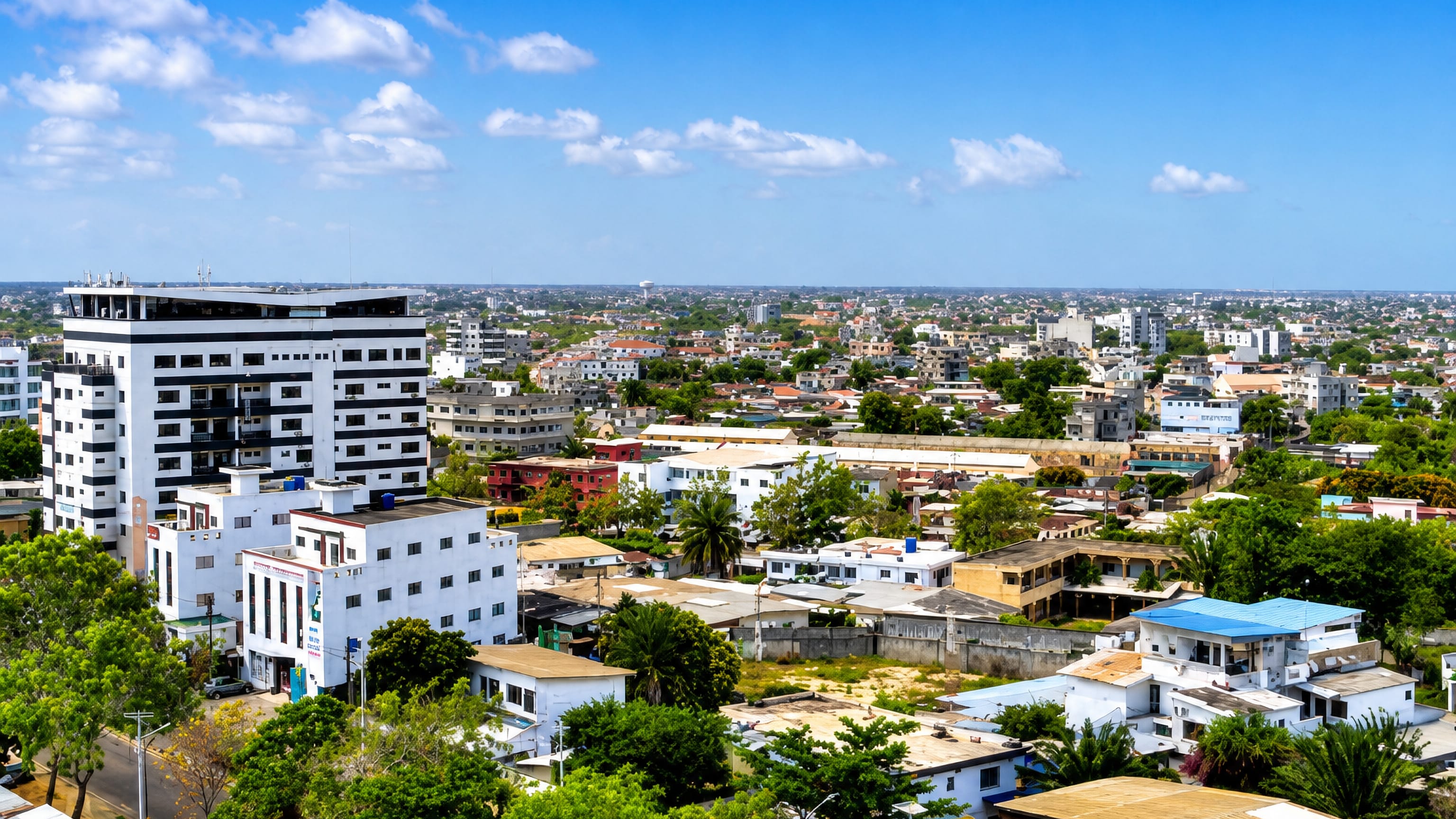 Aerial view over Cotonou, Benin, with white mid-rise blocks, tin-roofed houses and palms under a bright blue sky.