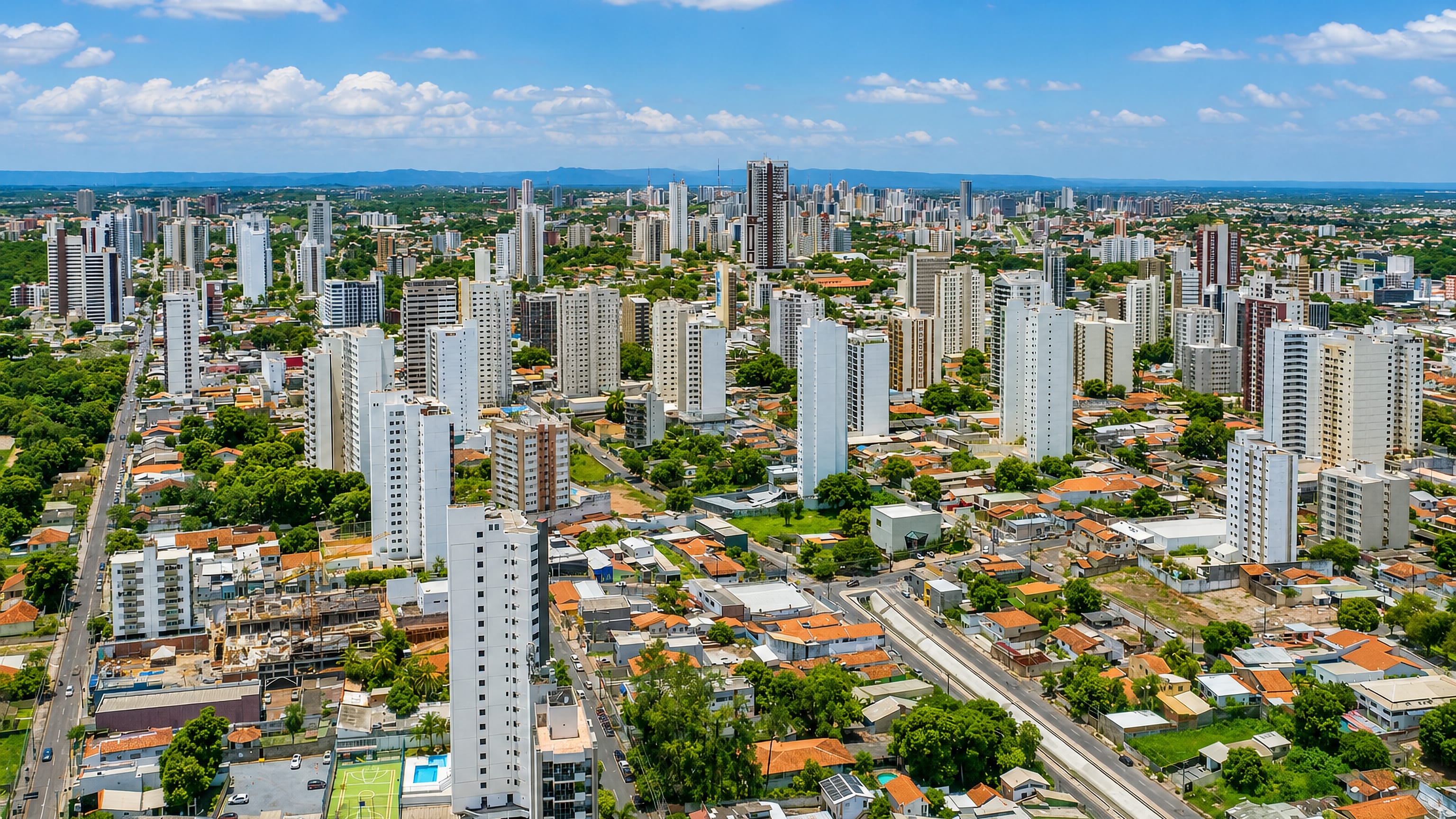 Aerial view of Cuiabá's apartment towers, red-tiled roofs and tree-lined avenues below distant hills.