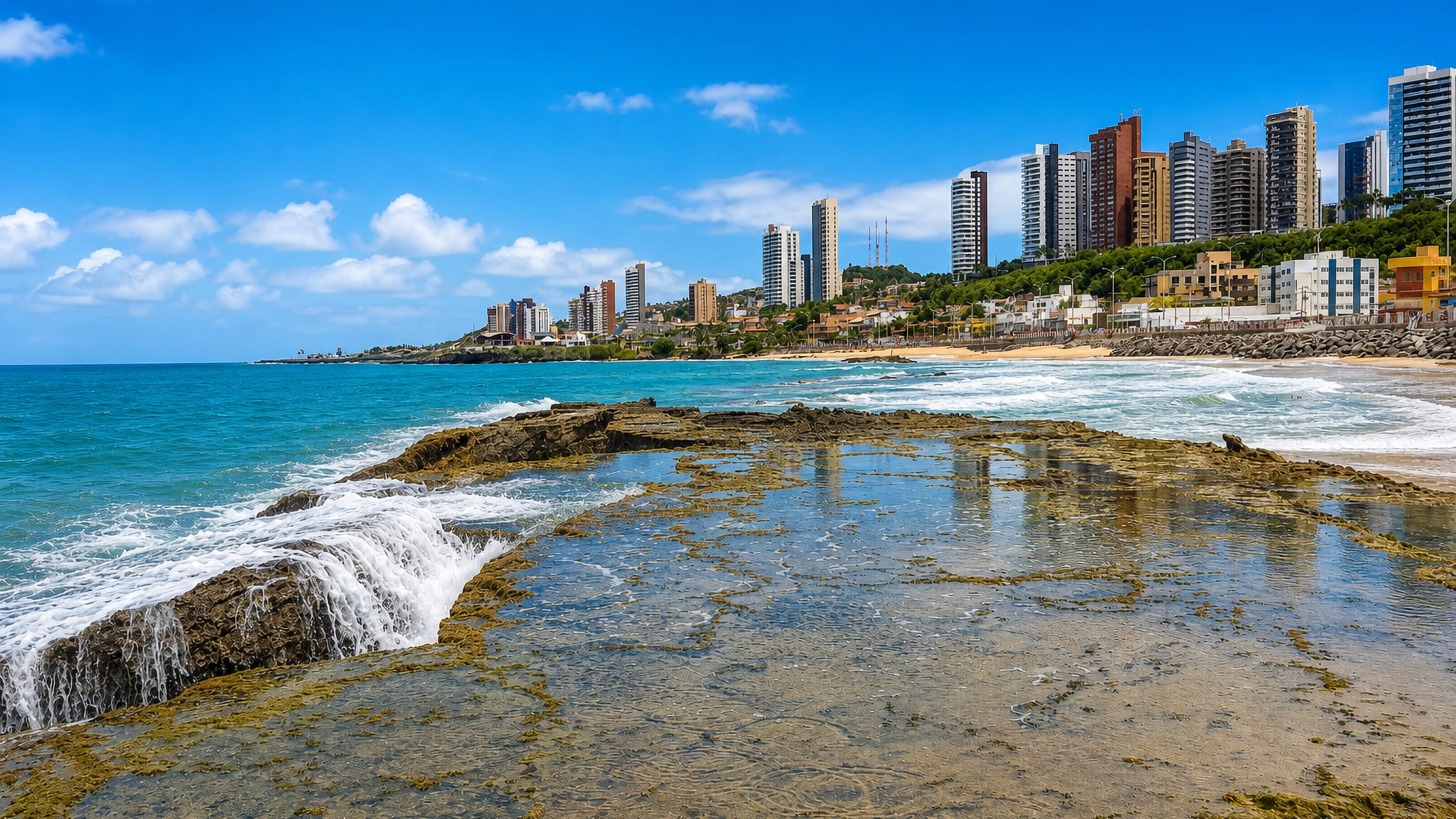 Rocky reef and surf at Praia do Meio, Natal, with beachfront tower blocks rising behind the sand.