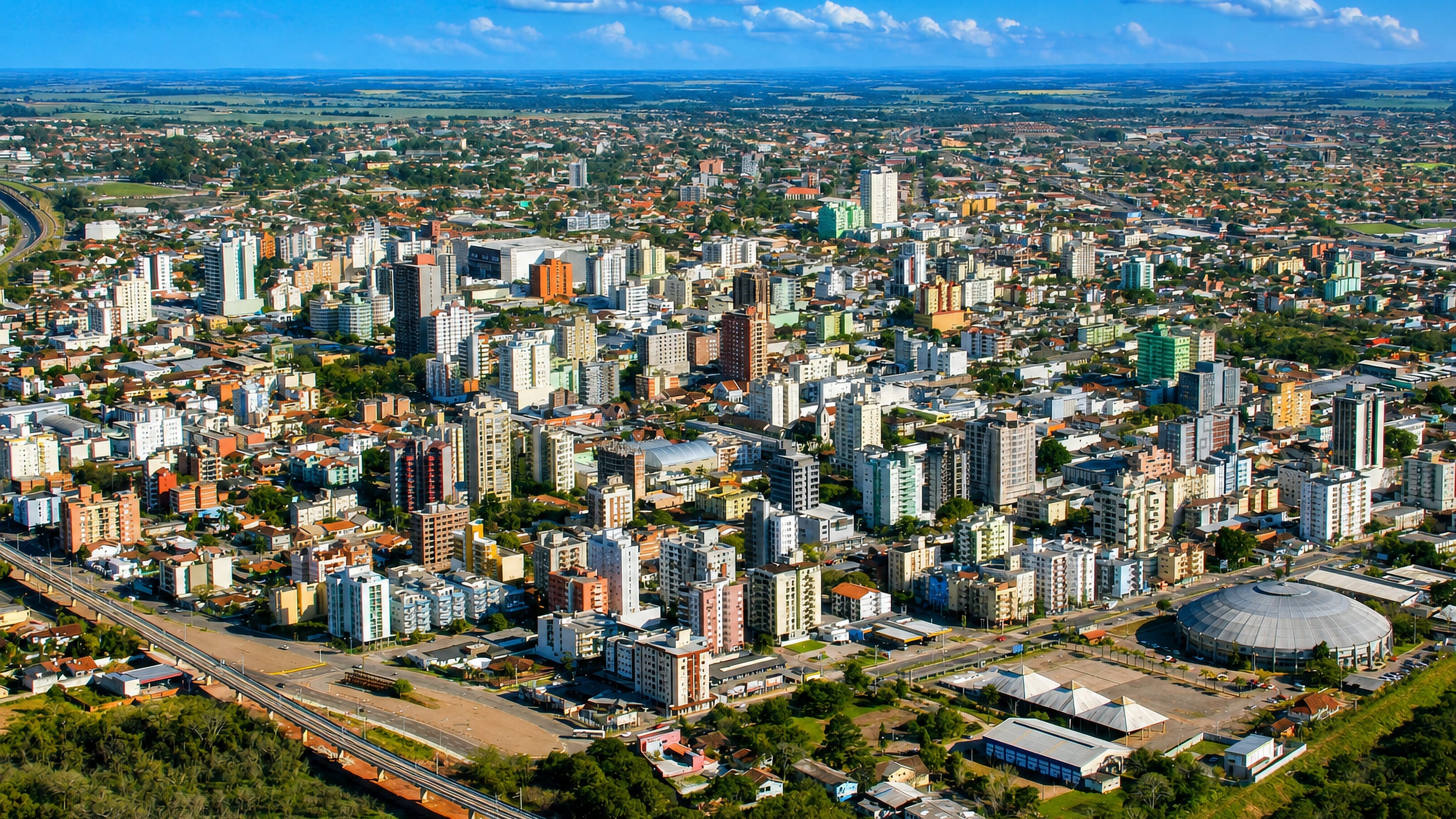 Aerial view of São Leopoldo, Rio Grande do Sul, with dense mid-rise blocks, a rail line and a domed arena.