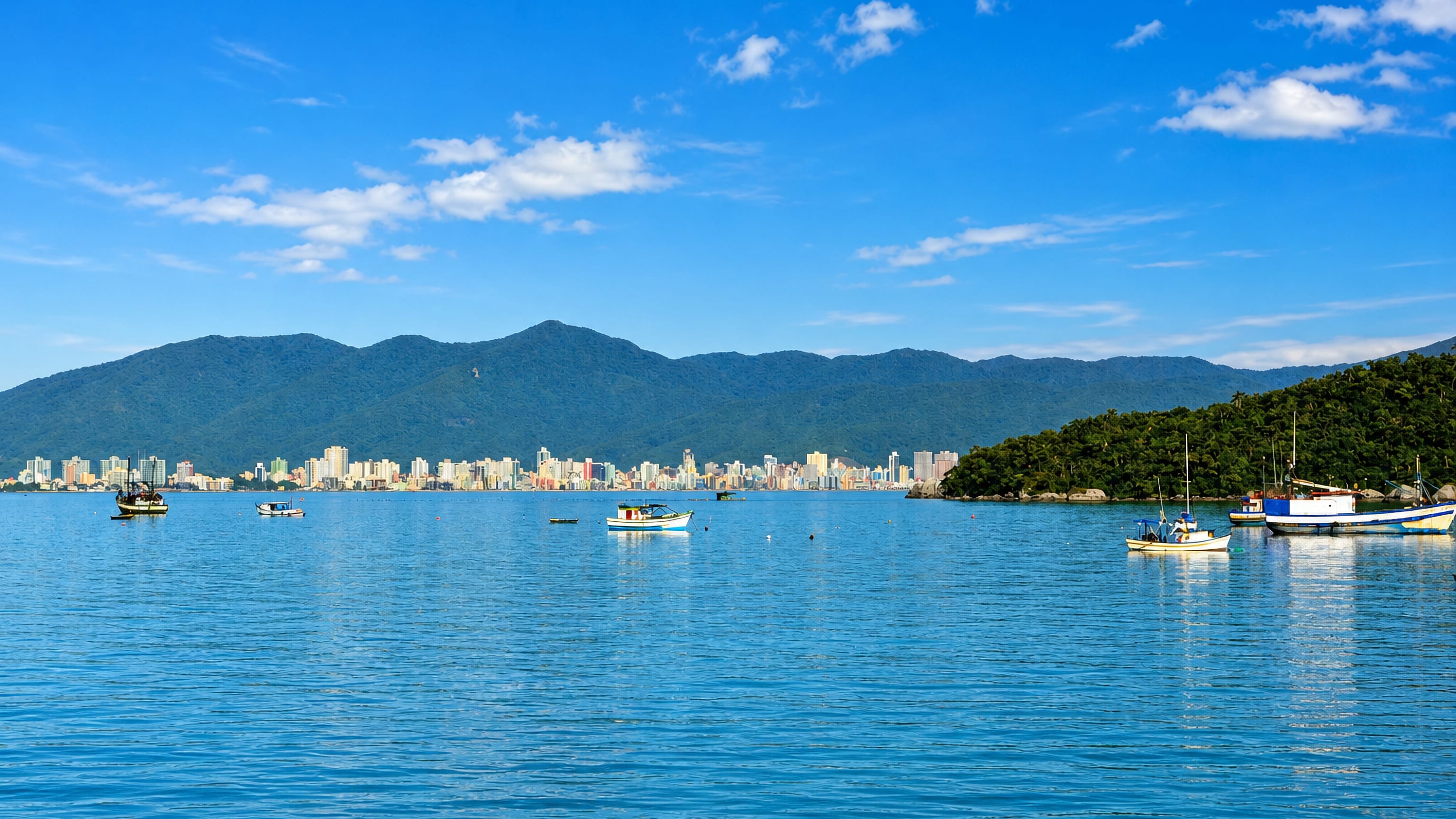 High-rise beachfront of Itapema, Brazil, across the bay beneath forested hills, with fishing boats moored.